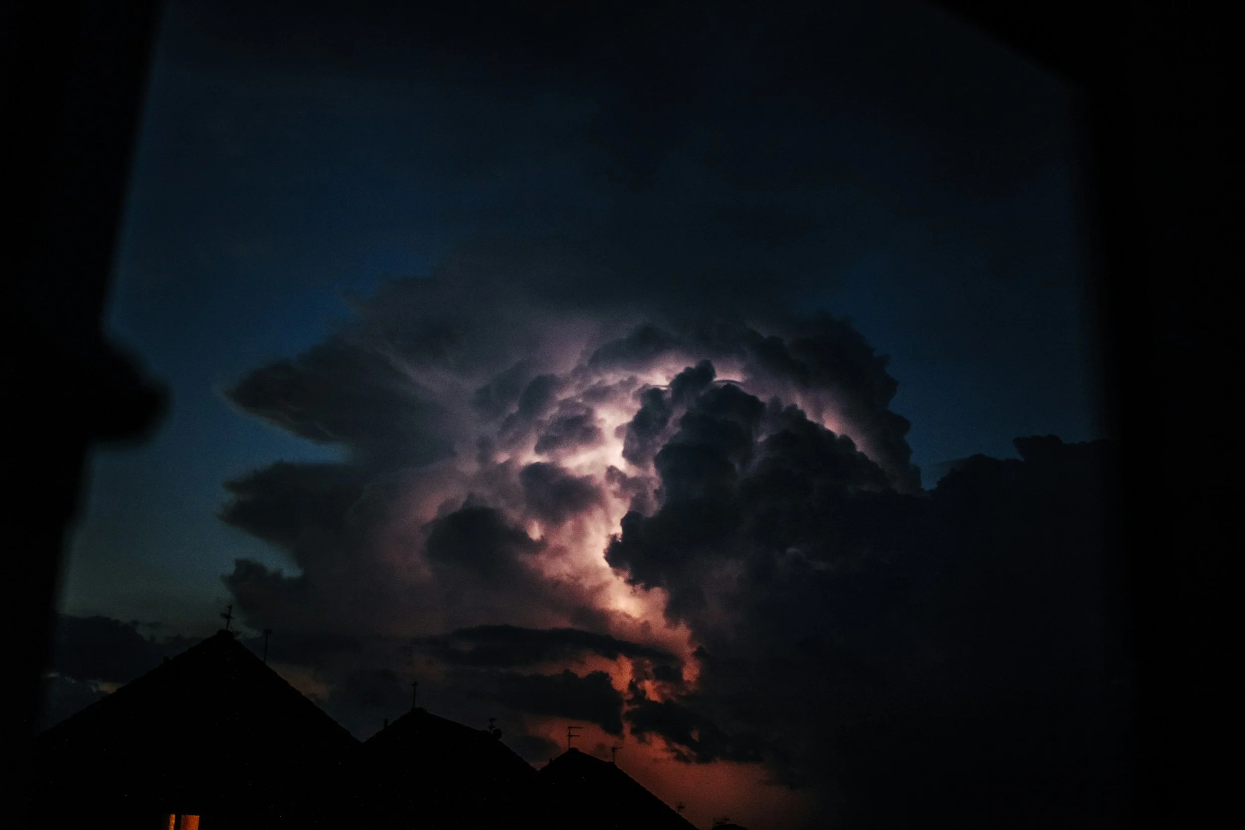 Dark storm clouds with lightning illuminating the clouds and a faint glow near the horizon, seen from a residential area at dusk.