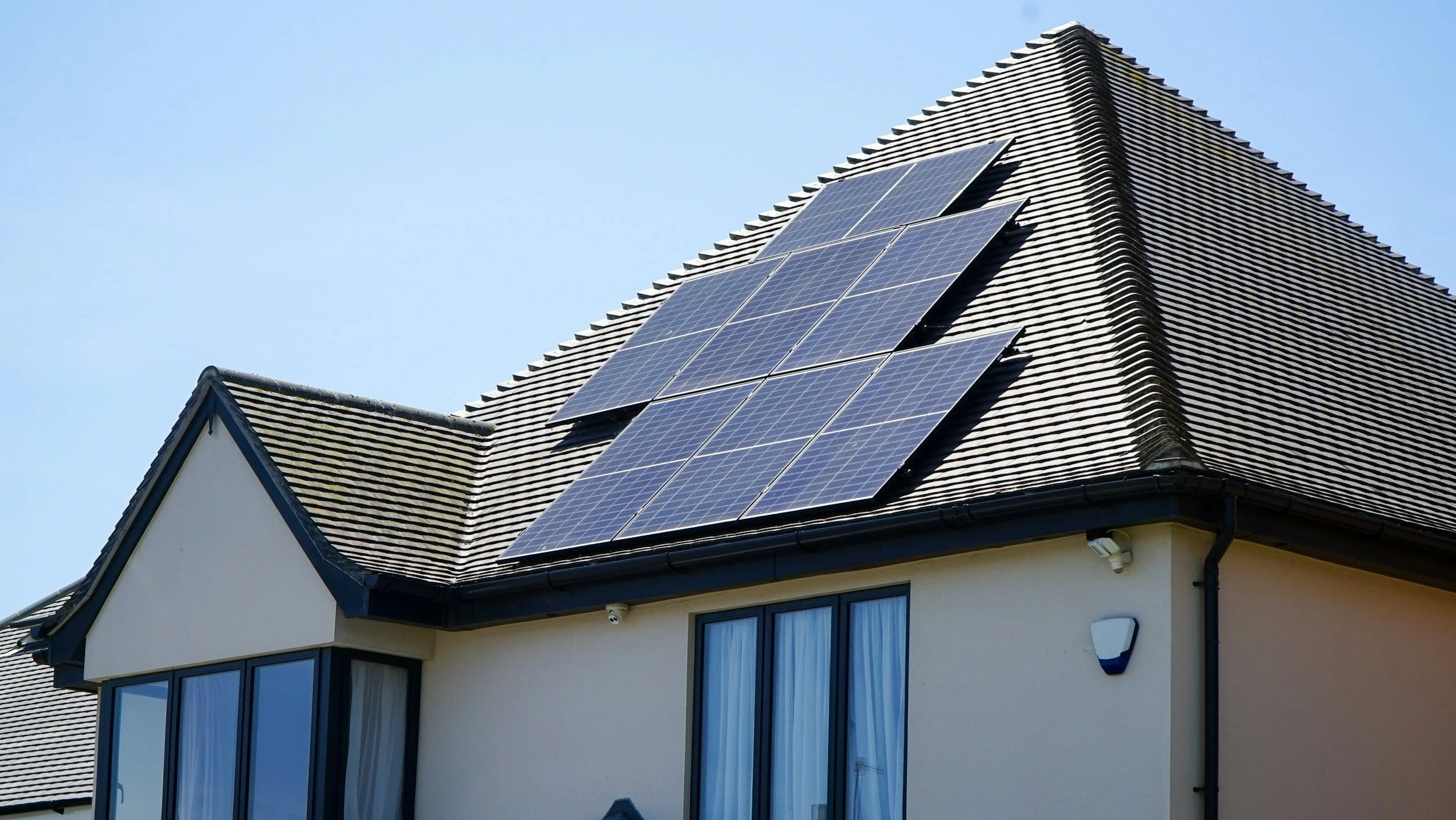 A house with a pitched roof fitted with multiple solar panels, located in a sunny environment against a clear blue sky.
