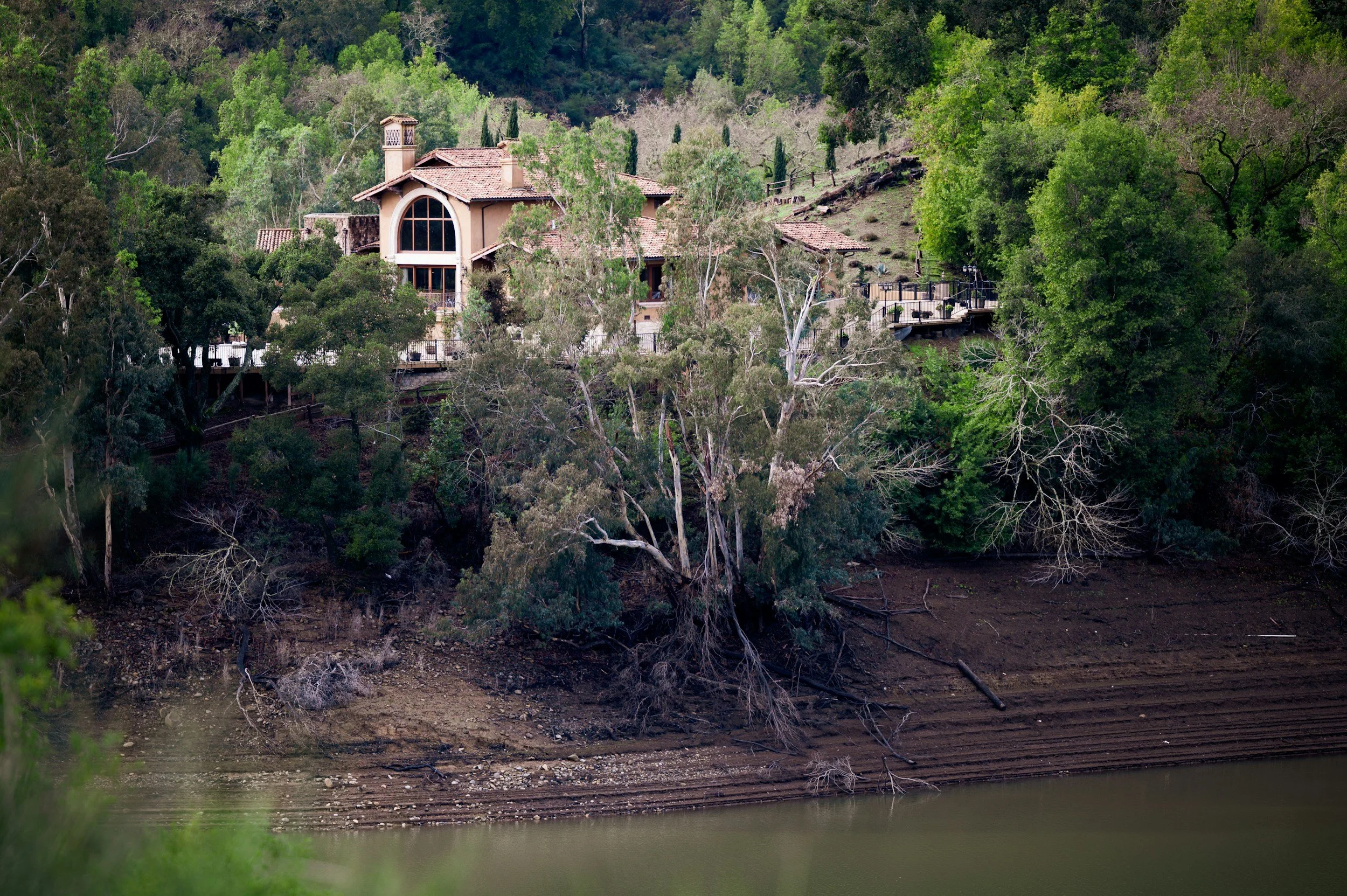 A house on a hillside surrounded by green trees, with the bottom of the image showing a body of water and a muddy shoreline.