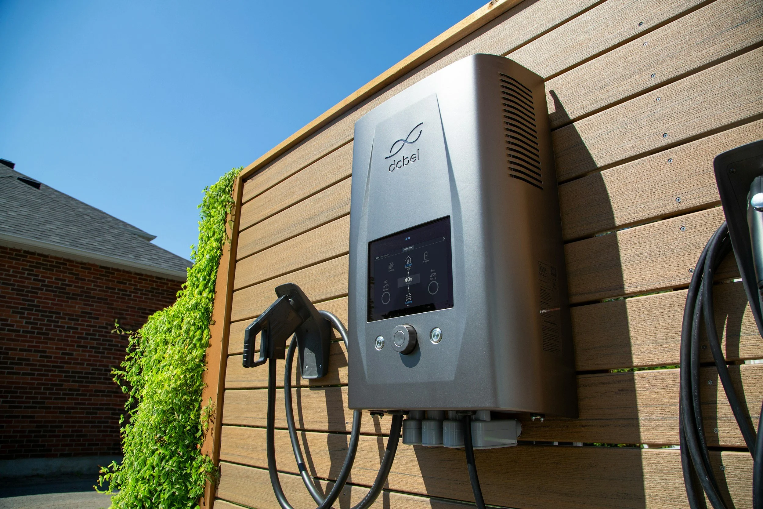 An electric vehicle charging station attached to a wooden wall outside a house, with a black charging cable connected to it.