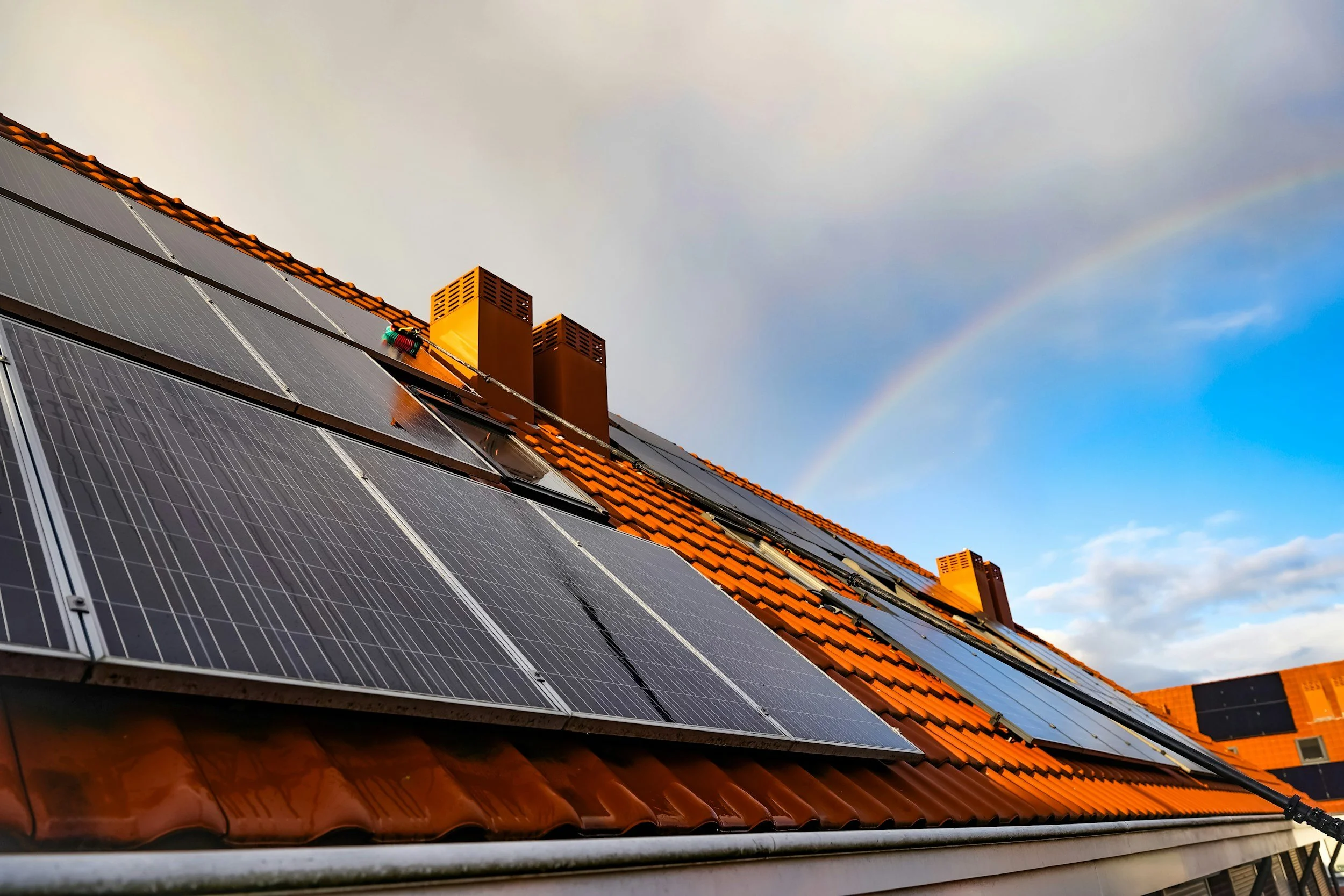 Solar panels installed on a rooftop with orange tiles under a sky with a faint rainbow.
