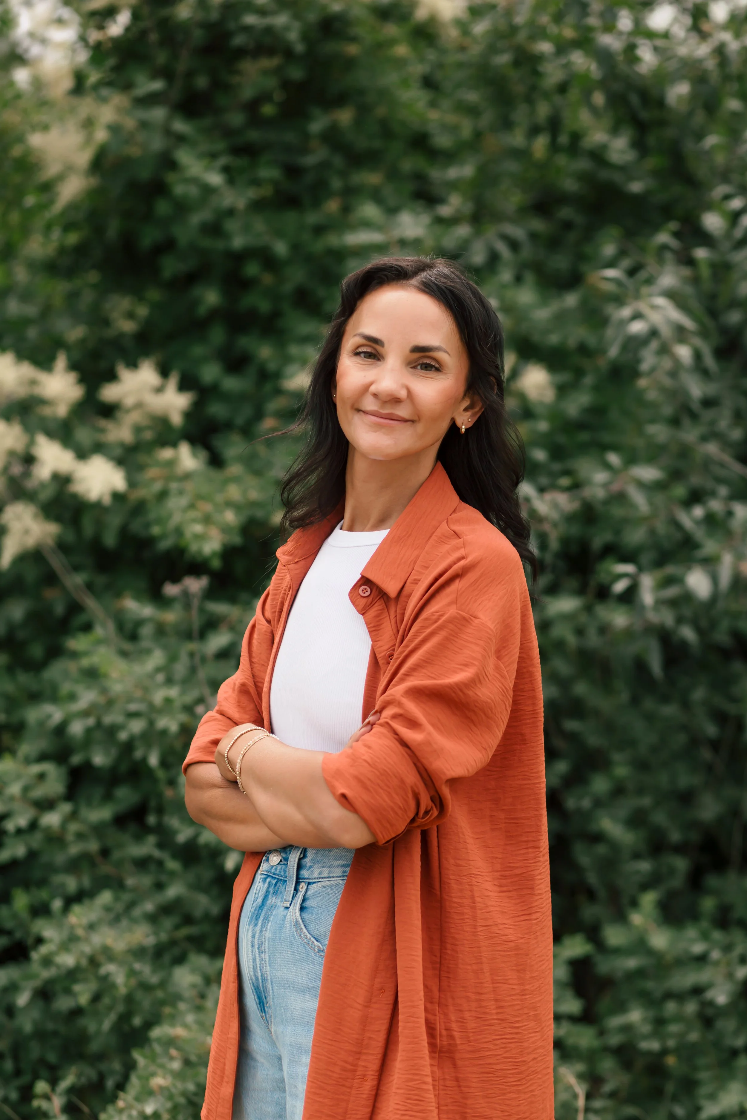 Psychedelic practitioner wearing a white shirt and orange jacket, smiling with arms crossed, standing outdoors with green foliage in the background.