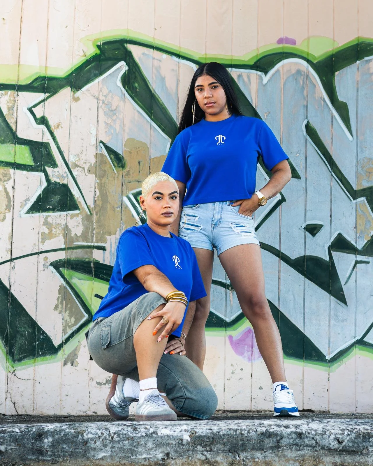 Two young women with dark hair and medium skin tones, wearing matching blue T-shirts with a white logo, posing in front of a graffiti mural on a wooden wall. One is kneeling with her hand on her knee, and the other is standing with one hand on her hip.