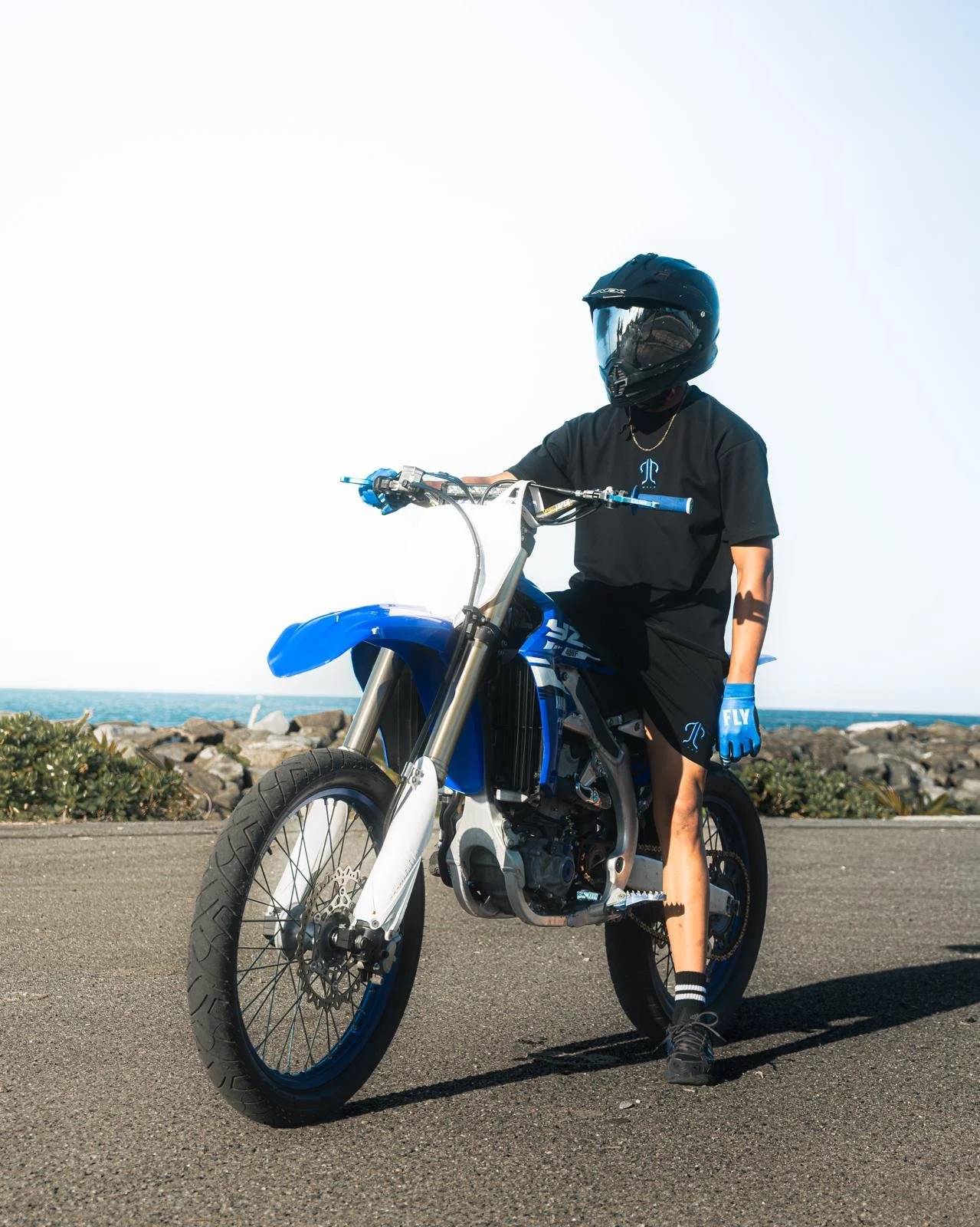 Person wearing a black helmet, black t-shirt, shorts, and blue gloves sitting on a blue and white dirt bike near the ocean with rocks and greenery in the background.