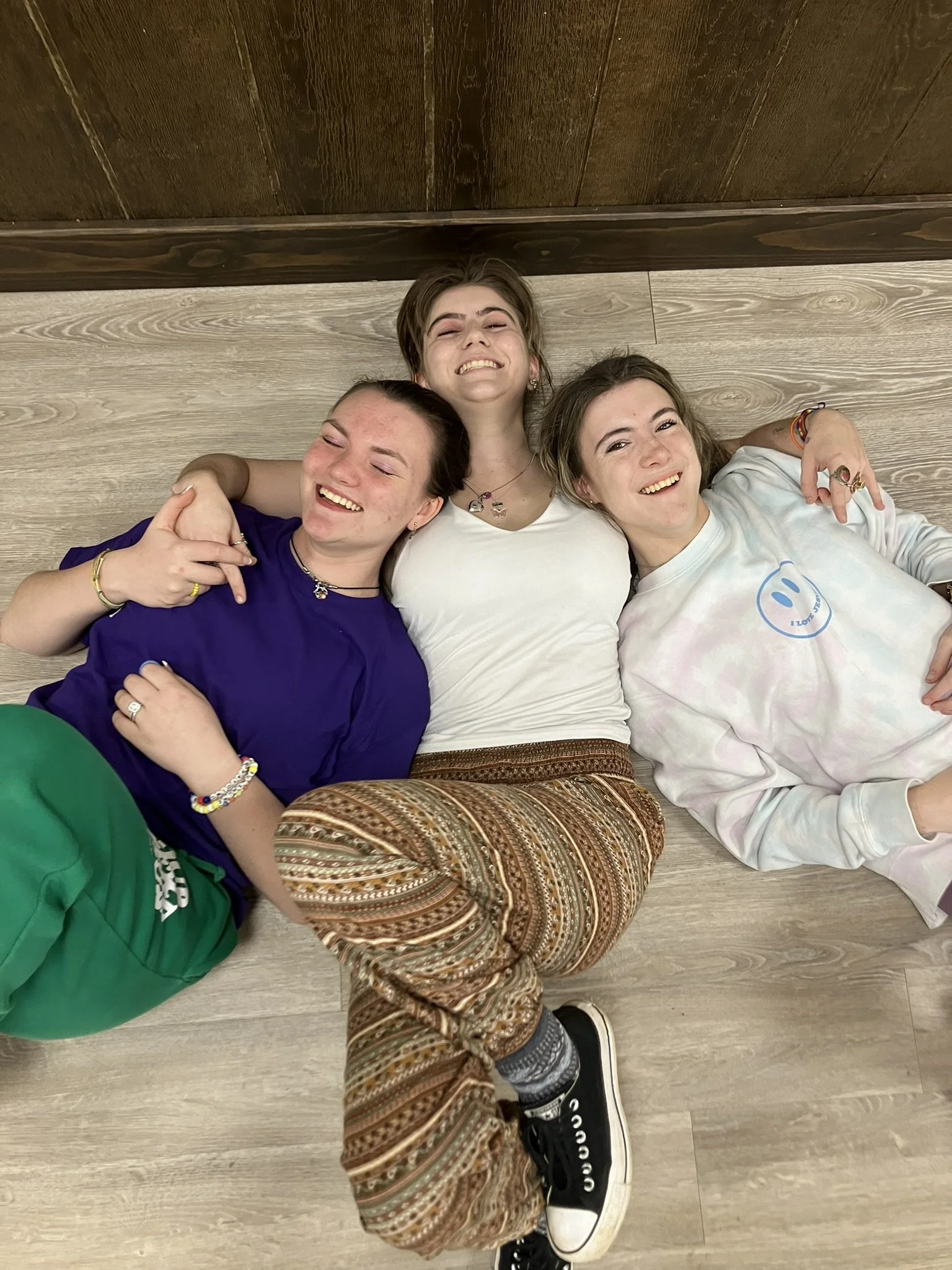 Three young people lying on the wooden floor, smiling and hugging each other.