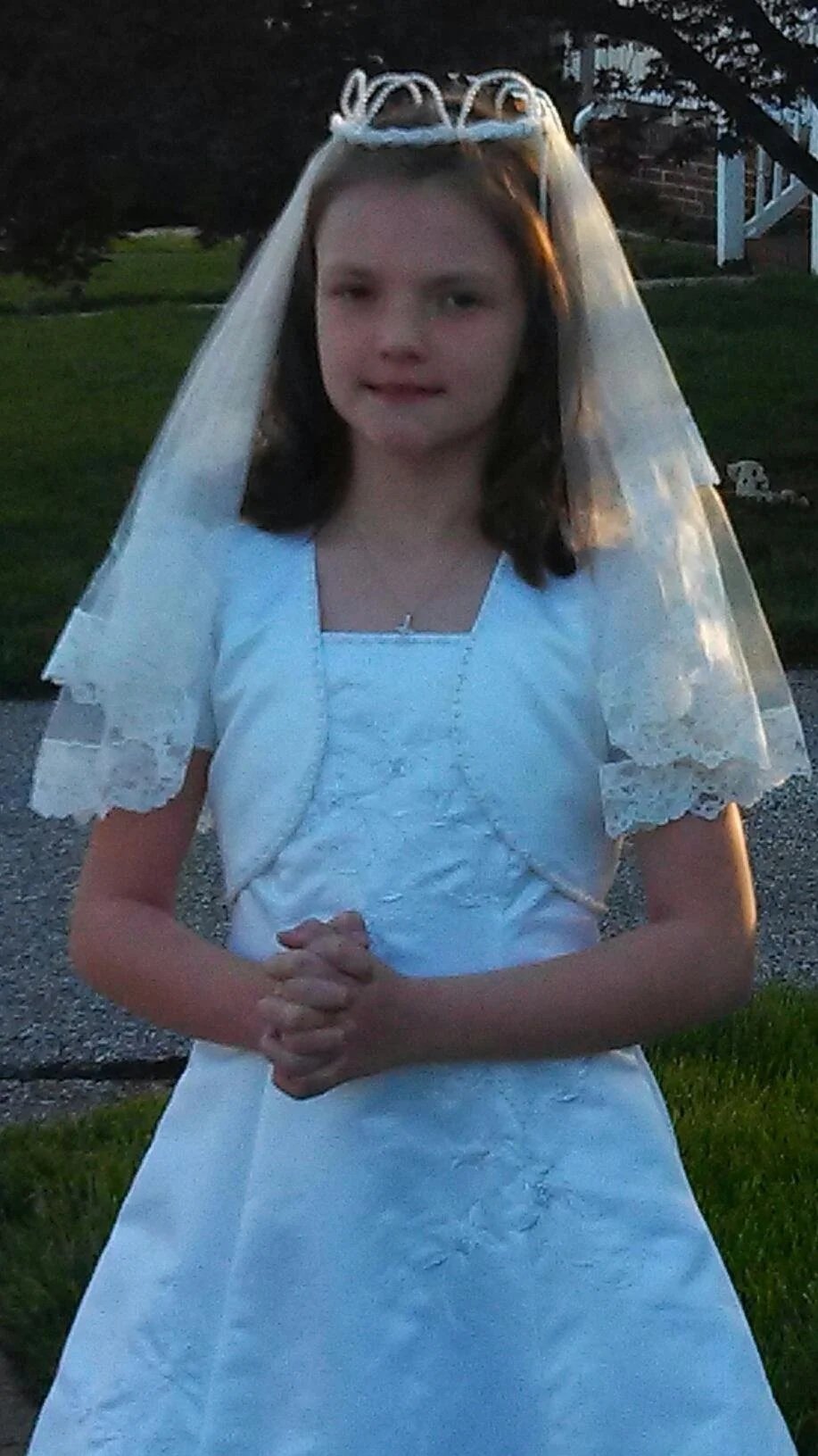 A young girl dressed in a white First Holy Communion dress with lace details, wearing a veil and tiara, standing outdoors at dusk with clasped hands.