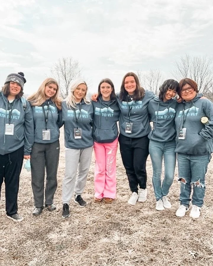 Group of seven women standing outdoors, wearing matching blue hoodies with text, posing with their arms around each other on a cloudy day.