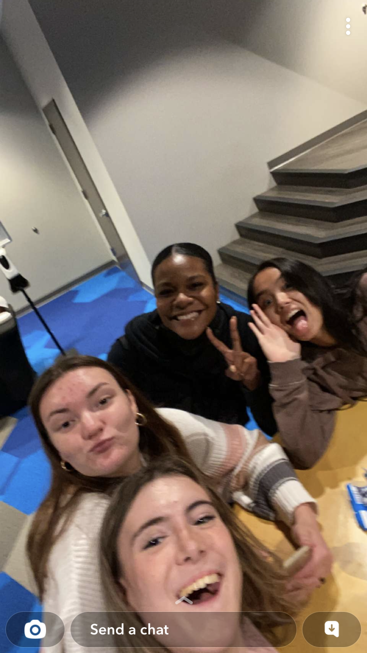 A group of five young women taking a selfie indoors, with happy and playful expressions, including sticking out tongues and making peace signs.