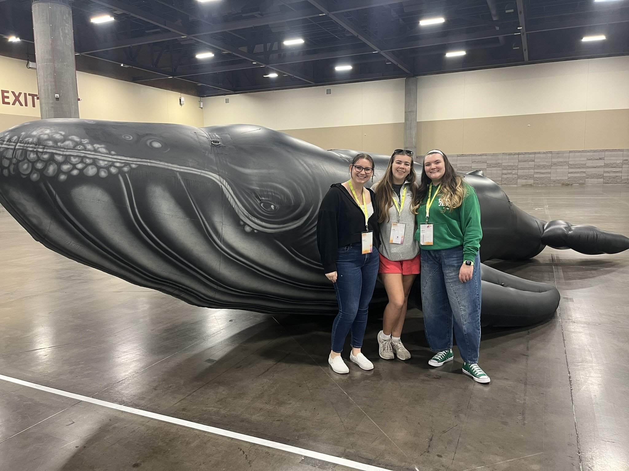 Three young women standing in front of a large whale sculpture inside a building with a high ceiling and concrete flooring.