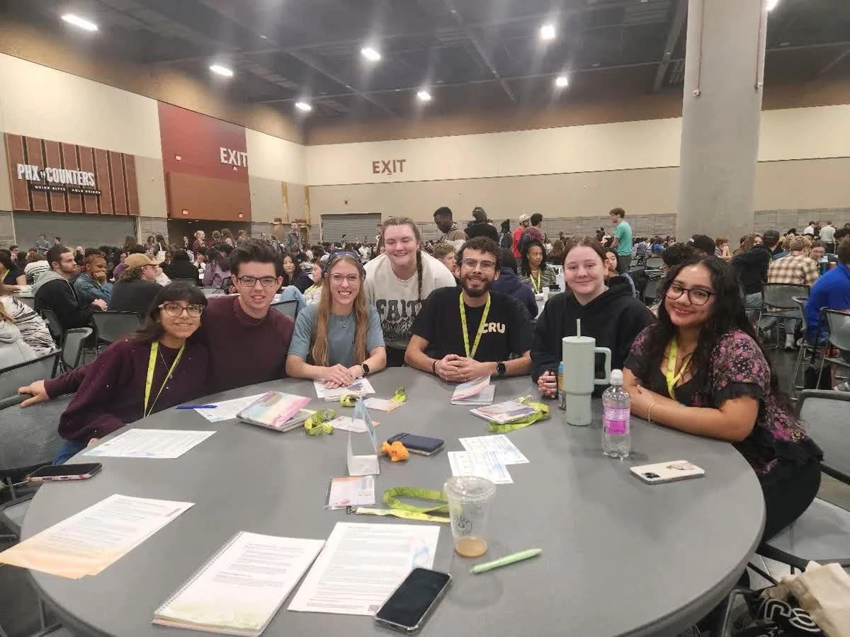 A group of seven young adults sitting around a table at a large indoor event or conference, with many people seated at tables in the background.
