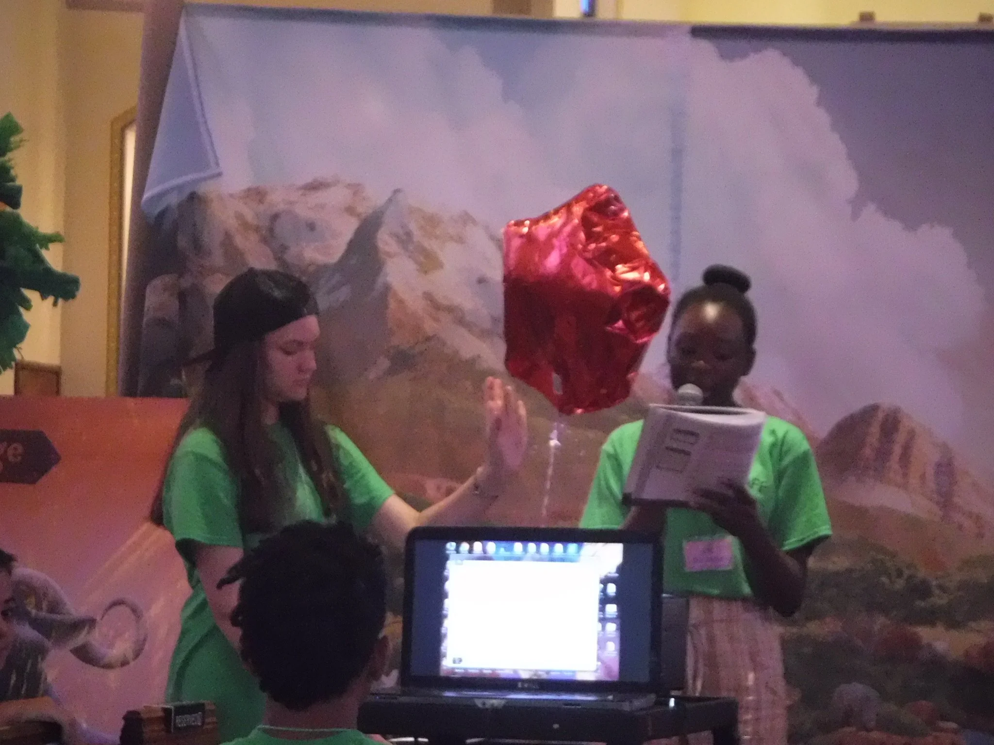 A group of people, some wearing bright green shirts, is gathered around a table or desk with a laptop. One person is holding a shiny red gift bag. Behind them, there is a backdrop with a mountain landscape and a cloudy sky. The setting appears to be an indoor event or gathering.