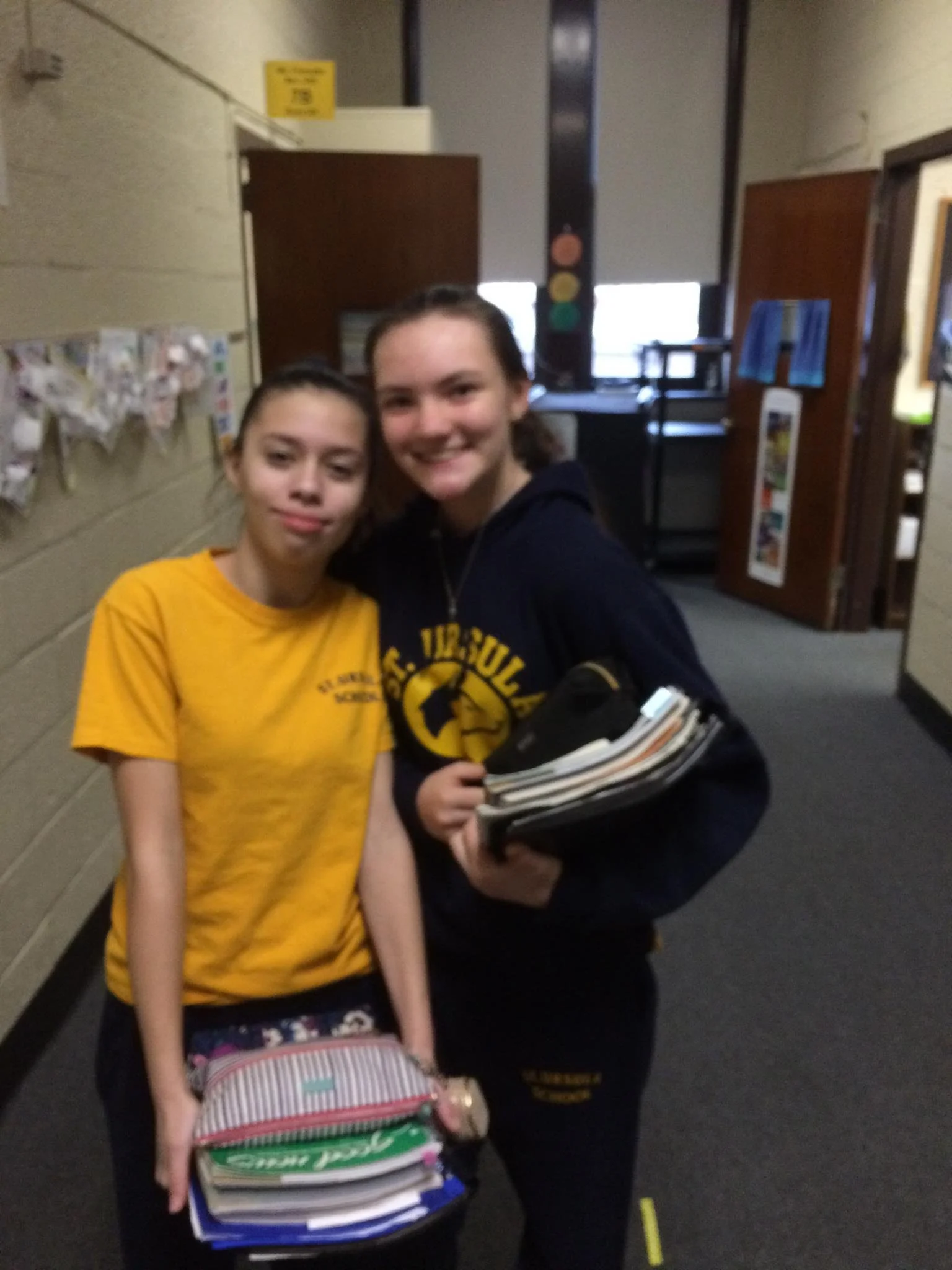 Two young girls standing indoors, smiling, with the girl on the right holding folders and notebooks, and the girl on the left holding a small bag or purse. The background includes a wall with some artwork and a door, with window blinds partially open.