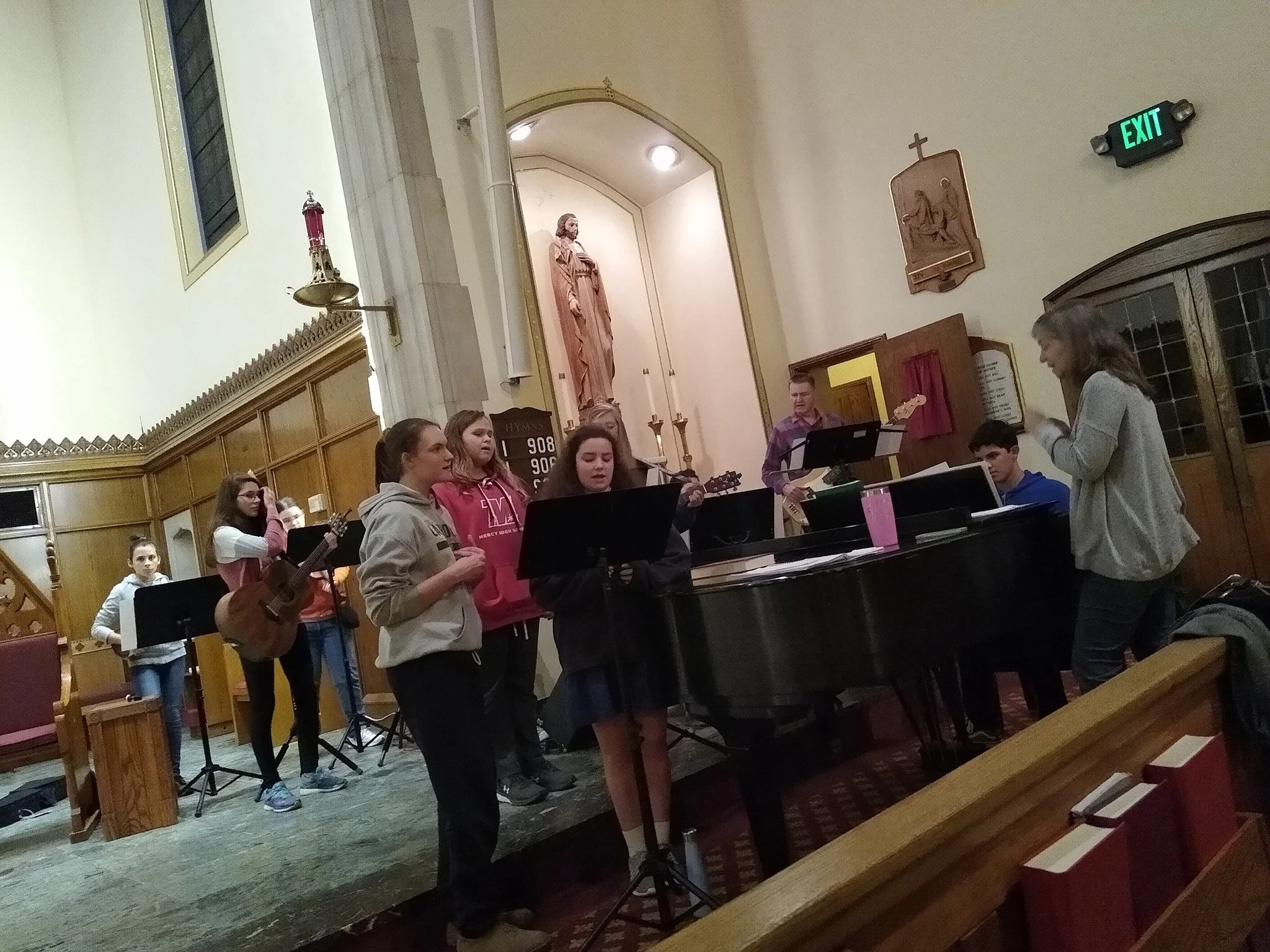 A group of young people singing and playing musical instruments in a church, with a religious statue and candle on the altar in the background.
