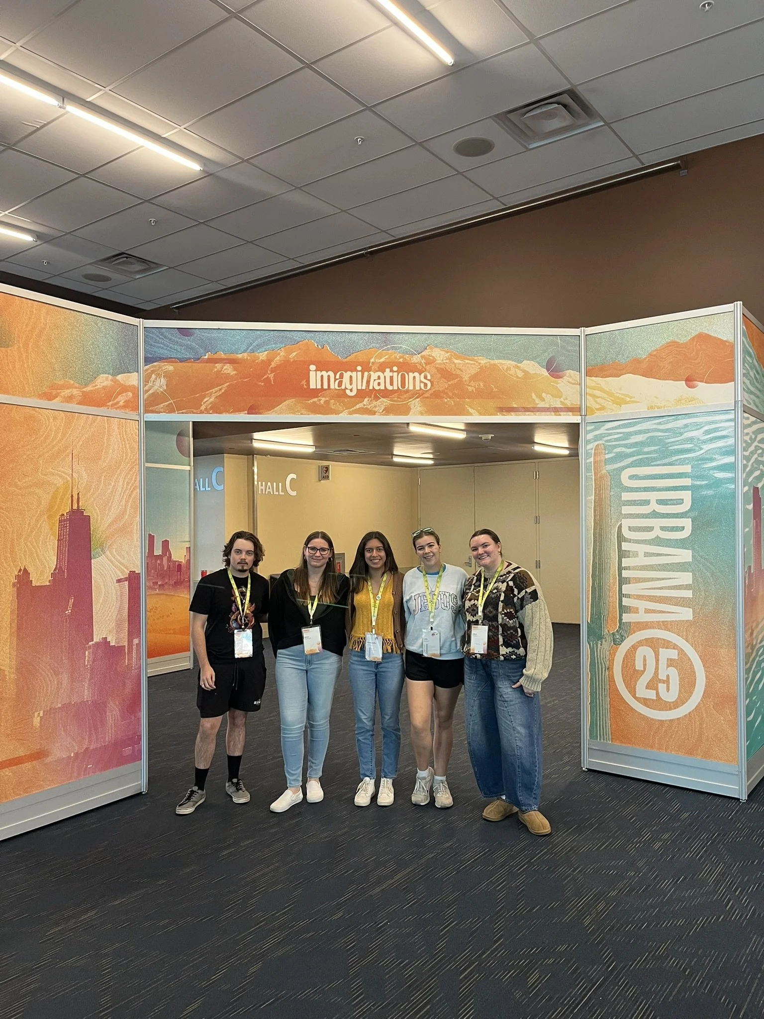 Five young women standing together indoors in front of a colorful display with cityscape and mountain graphics, smiling at the camera.