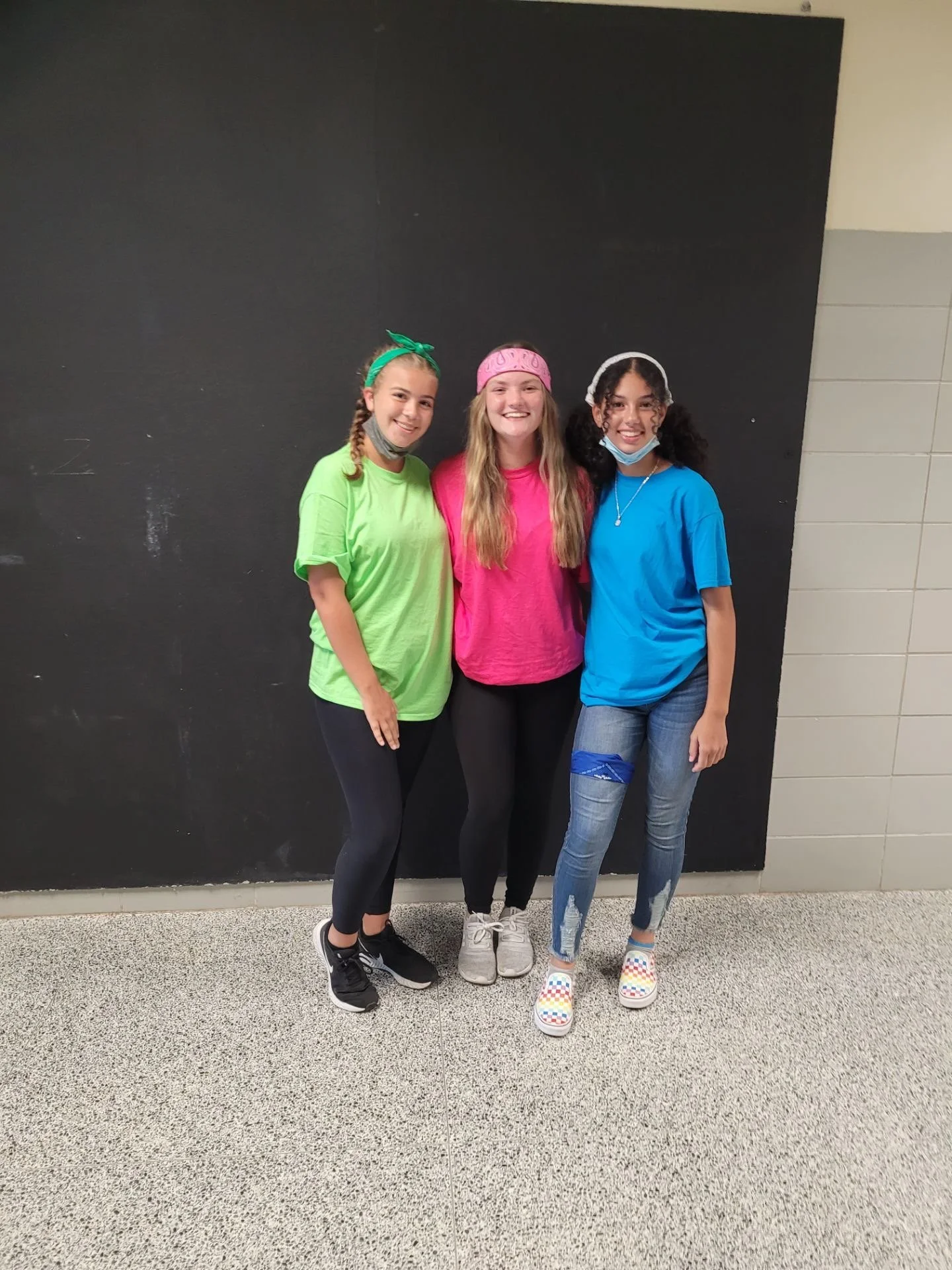 Three young girls standing together indoors in front of a black wall. They are smiling and wearing colorful casual clothing, including bright t-shirts and leggings, with headbands and face masks around their necks.