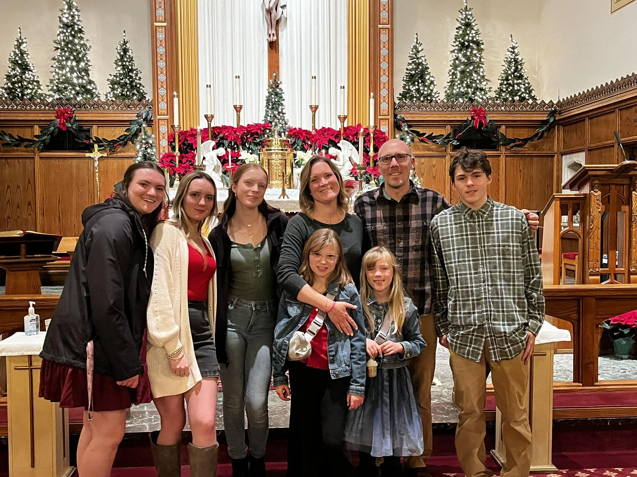 Family group posing in a church with Christmas decorations, including poinsettias, Christmas trees, and a crucifix, during holiday celebration.