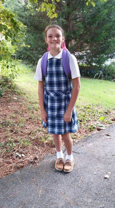 A young girl in a plaid school uniform with a white shirt and purple backpack standing on a paved path surrounded by greenery.