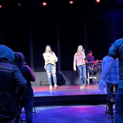 Two women singing on stage with microphone stands, with an audience watching, and a person playing keyboard in the background.