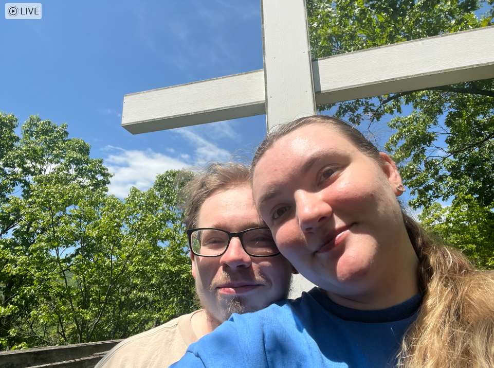 Two people taking a selfie outdoors in front of green trees and a large white cross, under a blue sky with some clouds.