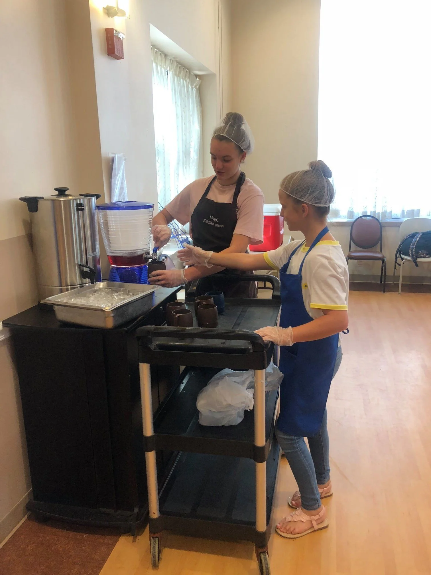 Two young girls wearing hair nets and gloves stand at a food service cart. One girl is filling cups with a red beverage from a beverage dispenser, while the other girl observes. The cart holds cups, a tray of ice, and other serving items. The room has light-colored walls, a window with curtains, and chairs against the wall.