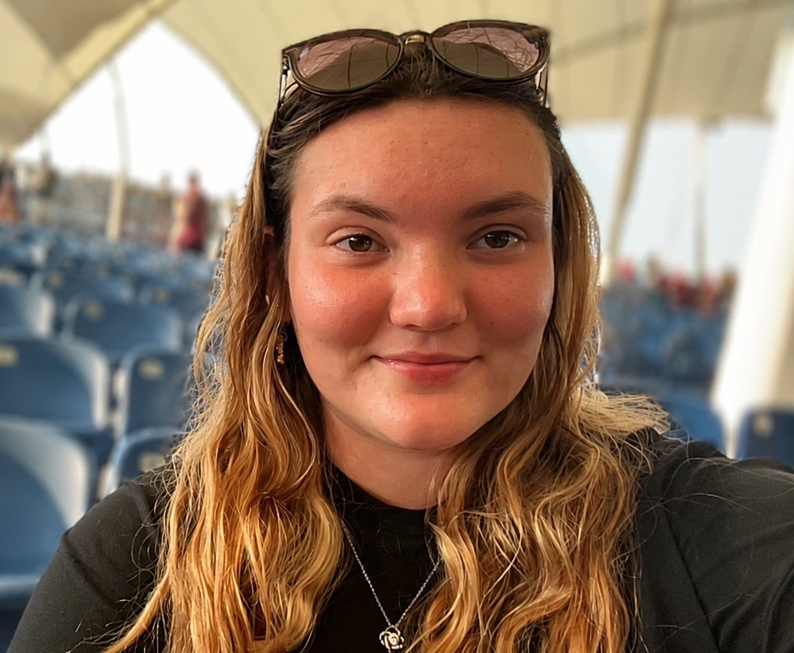 A young woman with long, wavy blonde hair, sunglasses on her head, smiling at the camera in an outdoor setting with blue chairs and a tent or canopy in the background.