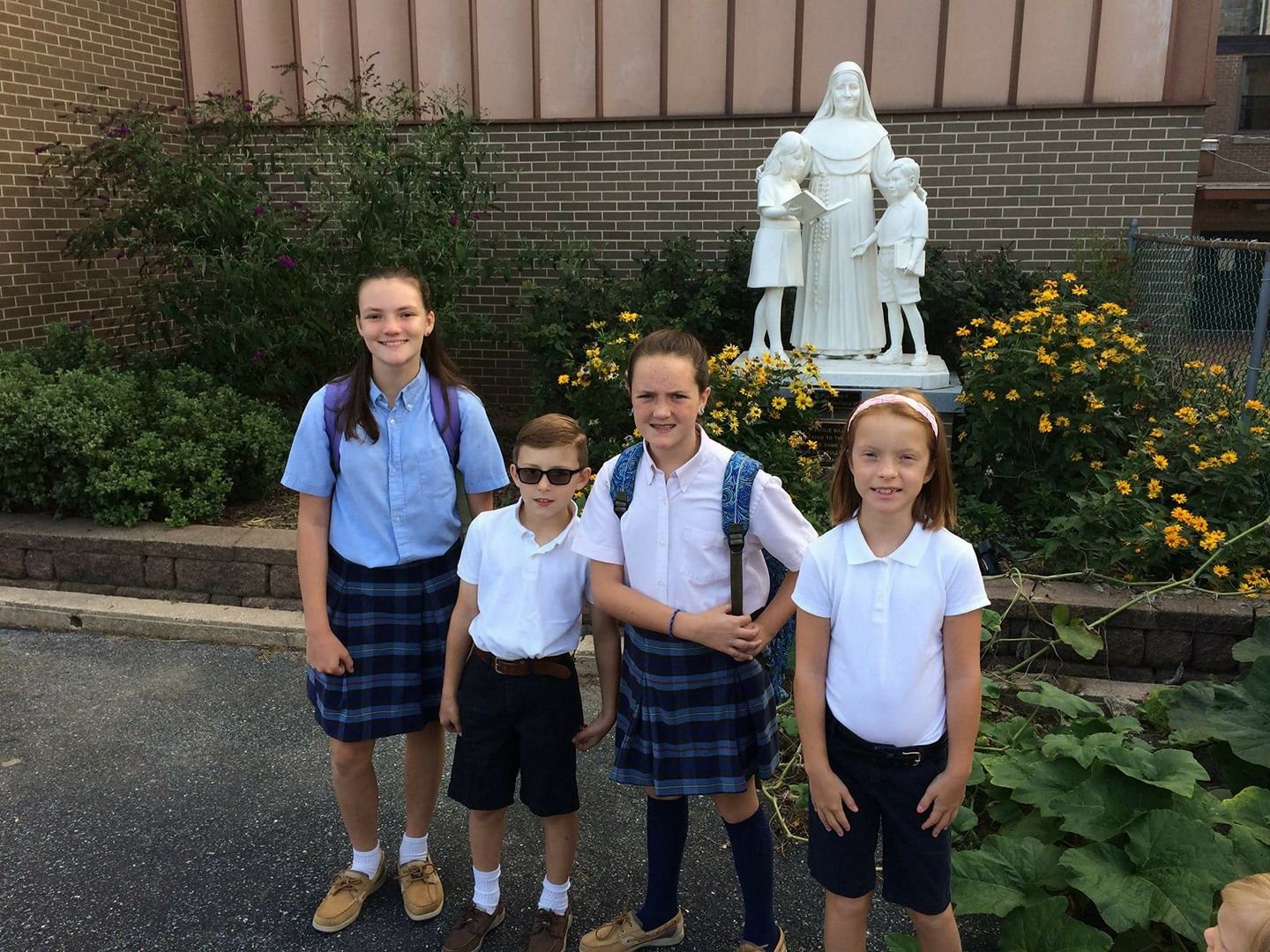 Four children in school uniforms standing outdoors in front of a religious statue, smiling at the camera.