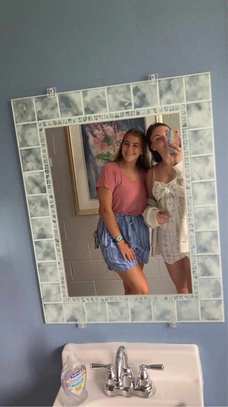 Two young women taking a selfie in a mirror in a bathroom. One woman is wearing a pink t-shirt and striped shorts, smiling. The other woman is wearing a white dress or cover-up, holding the phone. The bathroom sink and a soap dispenser are visible in the foreground, with a painting or picture hanging on the wall behind them.