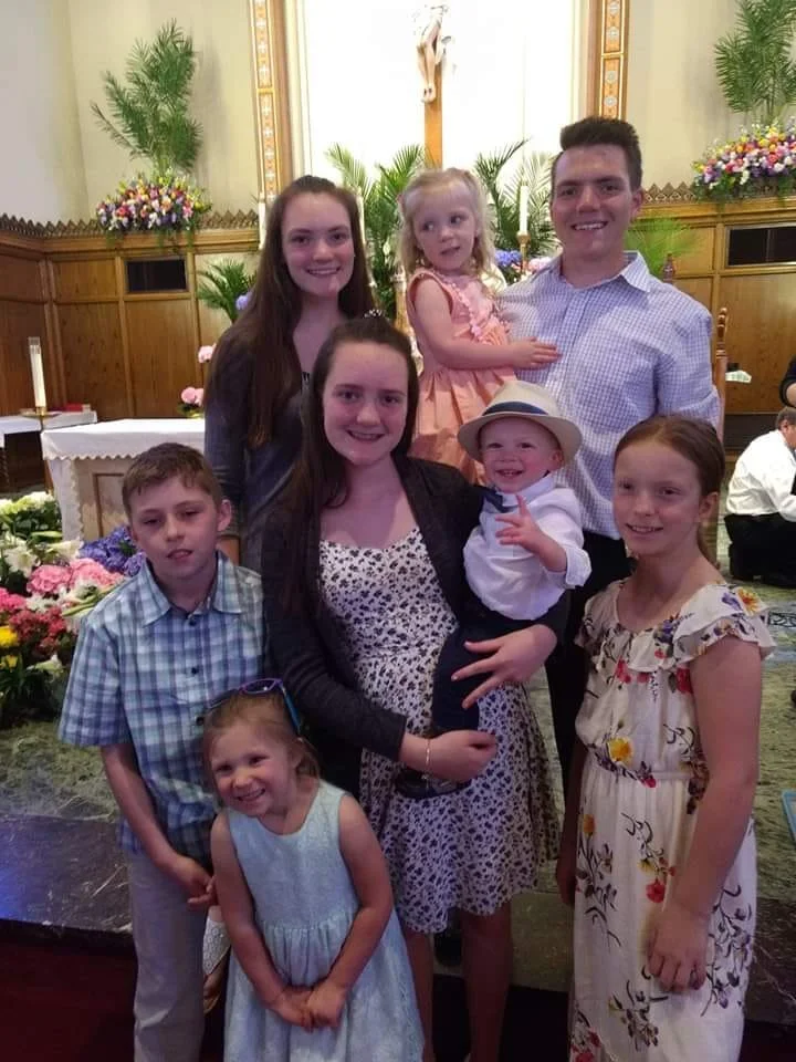 A family group of seven people, including children and adults, gathered inside a church or chapel with wooden paneling, flowers, and religious symbols, for a celebration or special occasion.