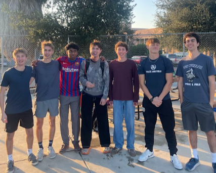 Group of seven teenage boys standing together outdoors on a sunny day, some wearing athletic clothing and backpacks, in a park or sports field area.