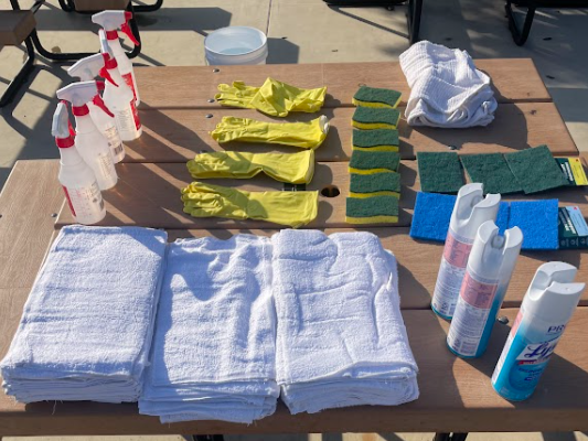Cleaning supplies and cloths arranged on a wooden table, including spray bottles, yellow rubber gloves, sponges, and folded towels.
