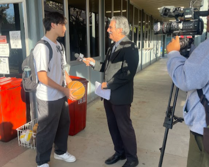 A news reporter interviewing a teenage boy with a basketball outside a store, with a camera operator filming the scene.