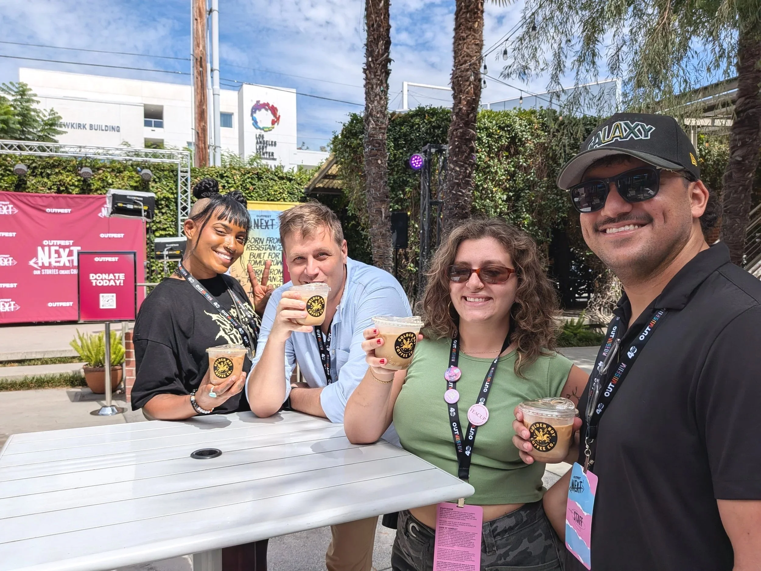 Group of five people smiling and holding coffee cups at an outdoor event. Four are women with one man, all wearing event lanyards, in front of a pink and white event sign and a stage with palm trees and a building with a large logo in the background.