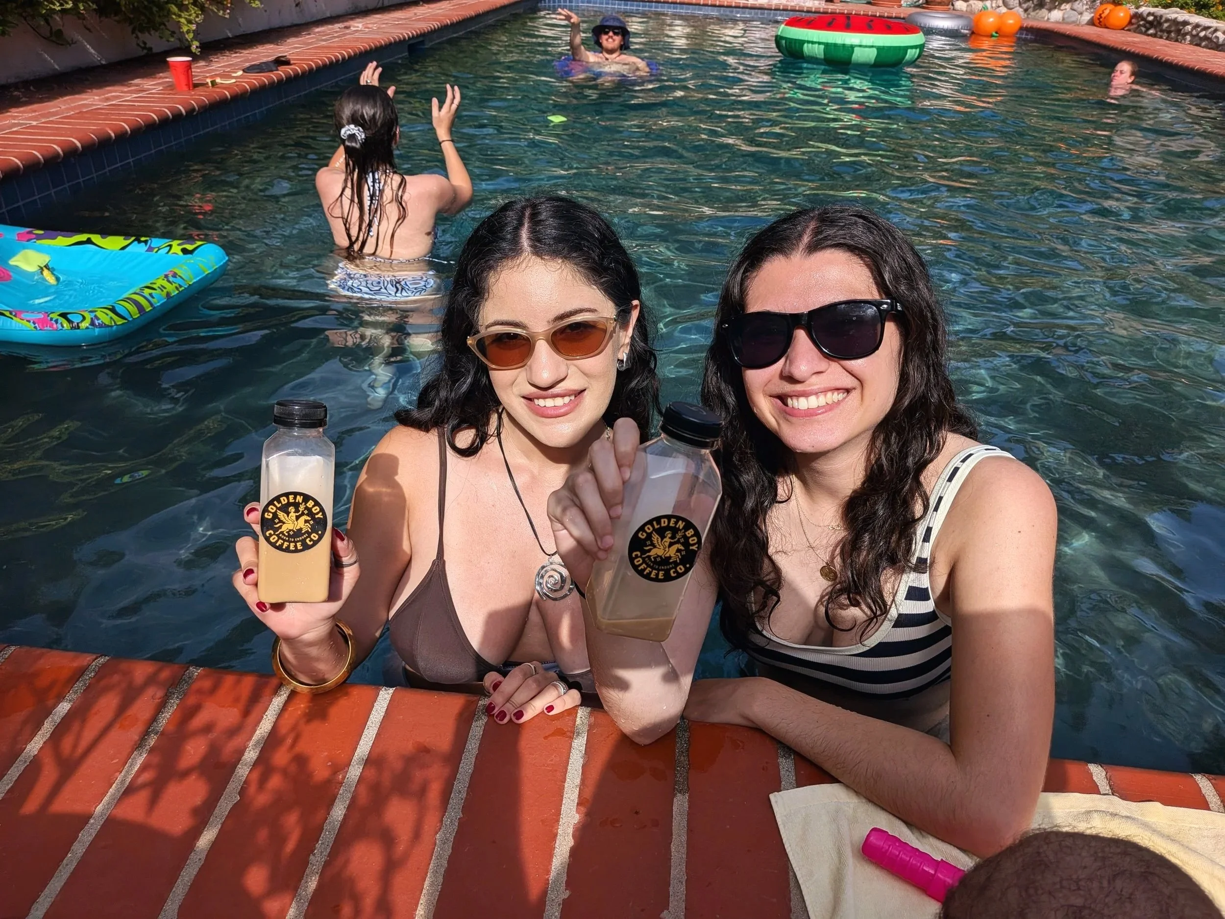 Two women are taking a selfie in a swimming pool. They are holding bottles of coffee labeled 'Golden Boy Coffee Co.' and smiling at the camera. There are people swimming and floating in the background, along with inflatable toys including a watermelo