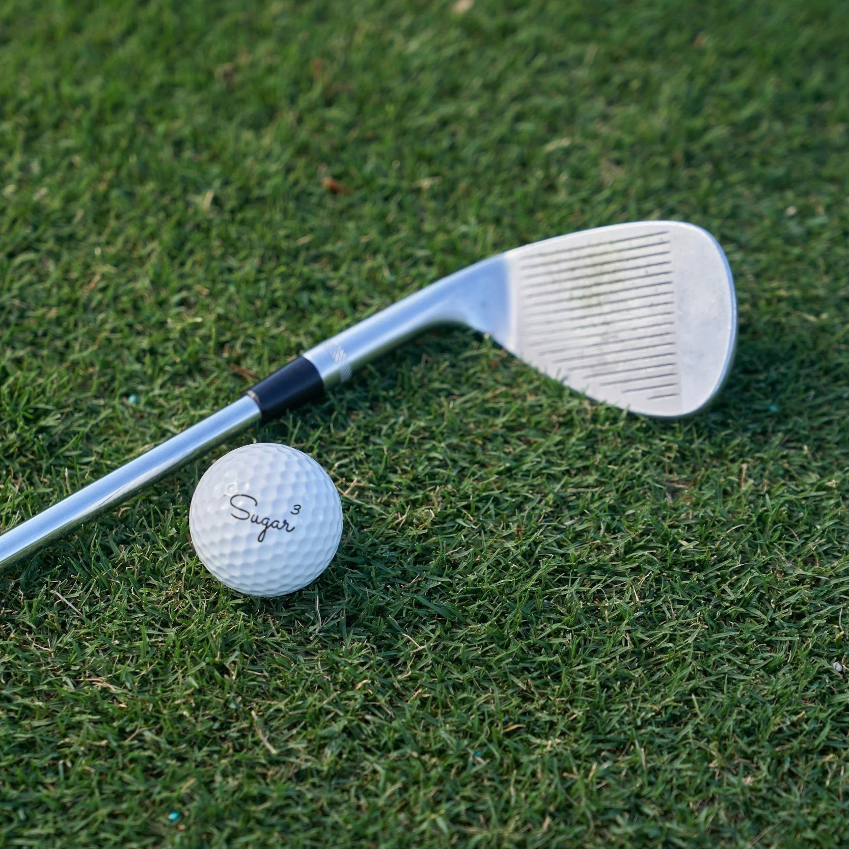 A golf club lying on green grass next to a golf ball with the word 'Sugar' written on it.