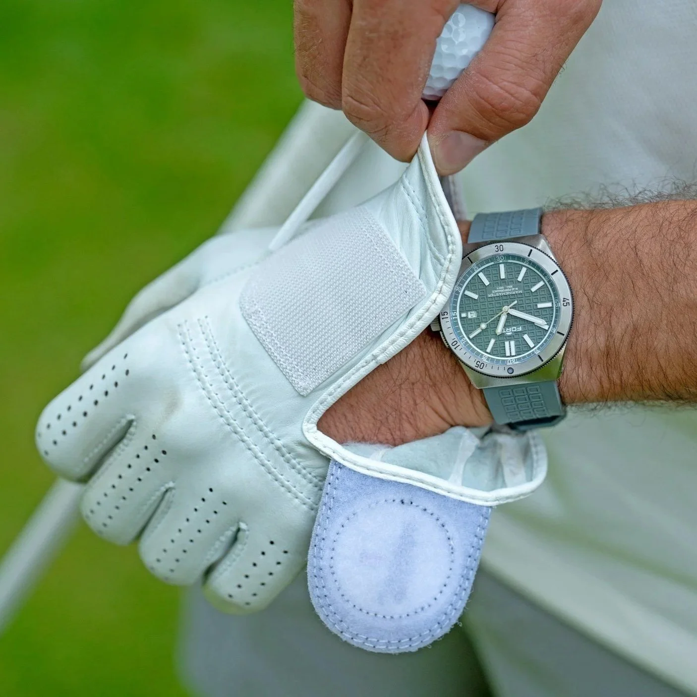 A golfer wearing a white golf glove and a gray wristwatch adjusting his glove on a golf course with a green background.