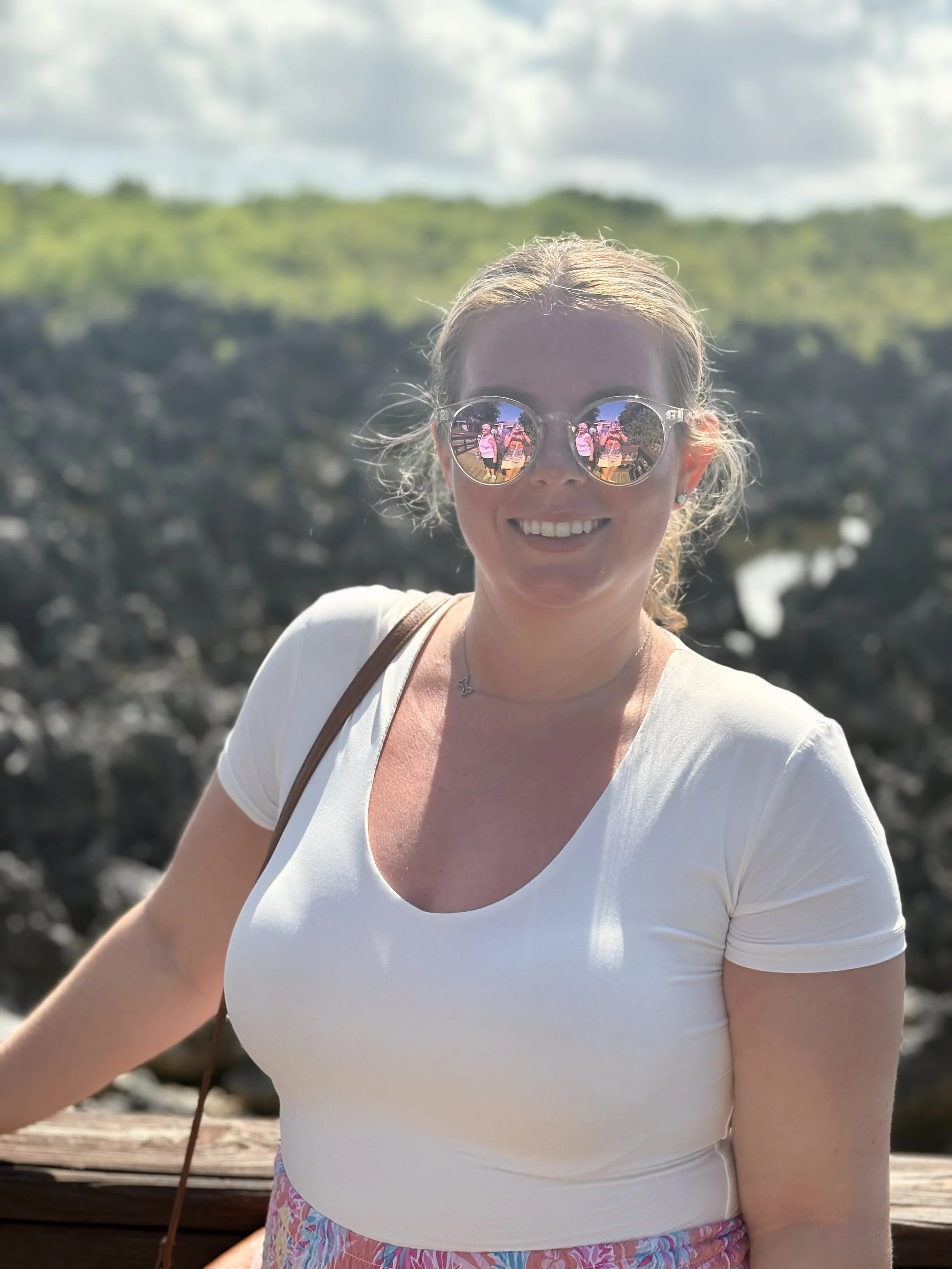 A smiling woman with blonde hair and sunglasses reflecting a person taking her photo, standing outdoors with trees and cloudy sky in the background.