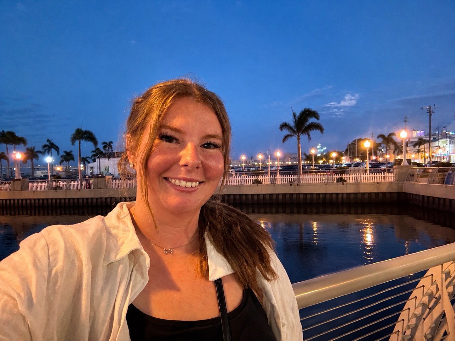 A smiling woman taking a selfie outdoors at dusk by a waterside with palm trees, lit street lamps, and a lively urban area in the background.