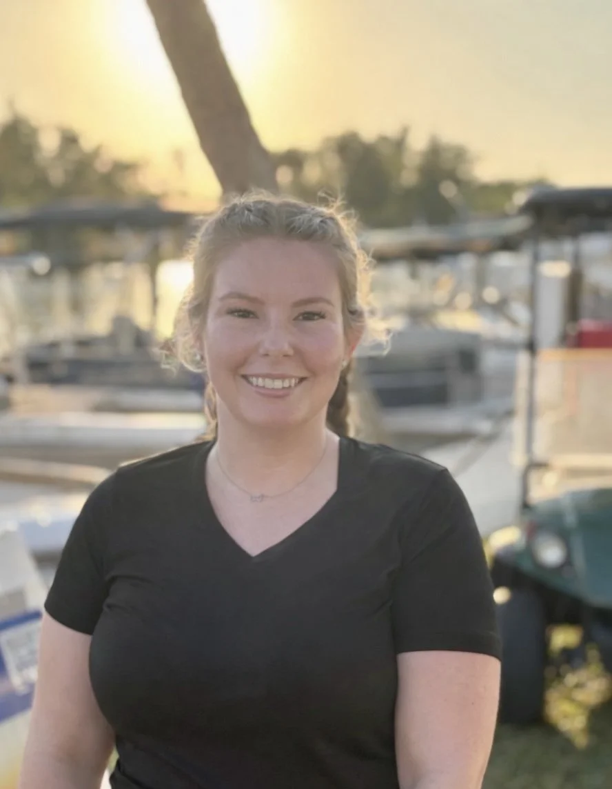 A young woman smiling outdoors with boats and a tree in the background during sunset.
