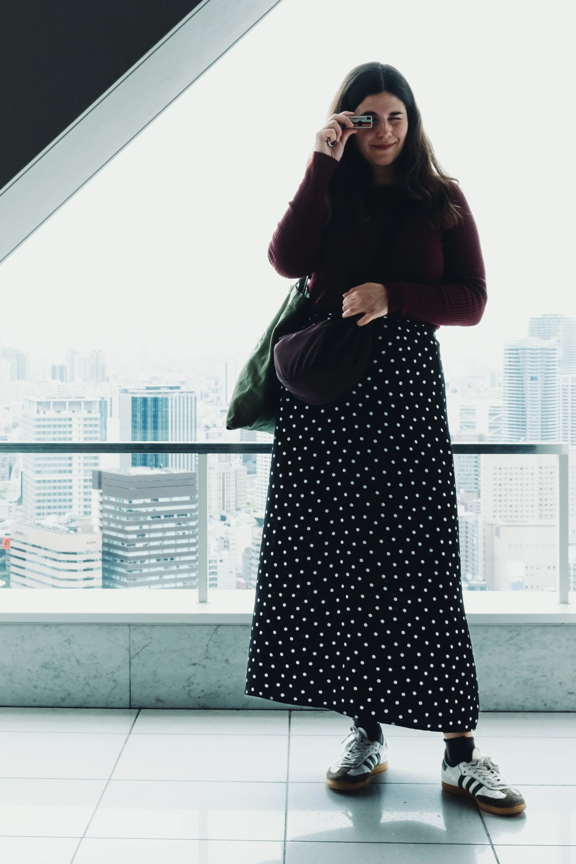 Young woman wearing a scurt, sneakers, and a maroon top standing inside a high-rise building with large windows, city skyline view in the background, holding a camera to her eye with a playful expression.