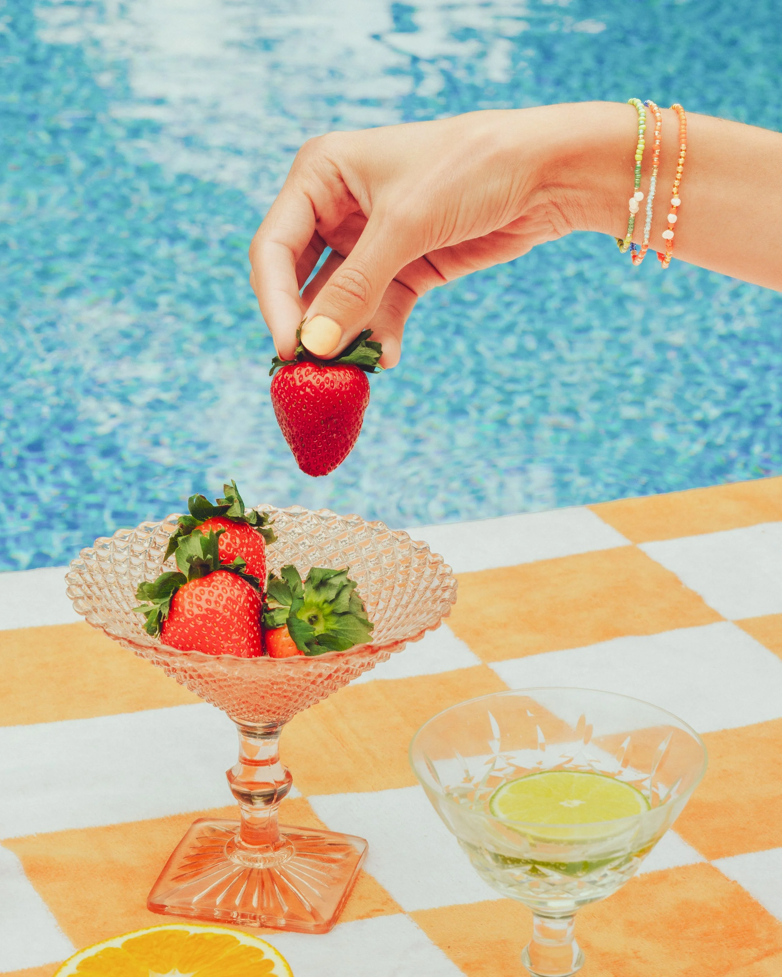 A person's hand dropping a strawberry into a pink glass bowl filled with strawberries. The scene is outdoors near a swimming pool, with orange and white checkered tablecloth and a glass bowl of lime slices nearby.