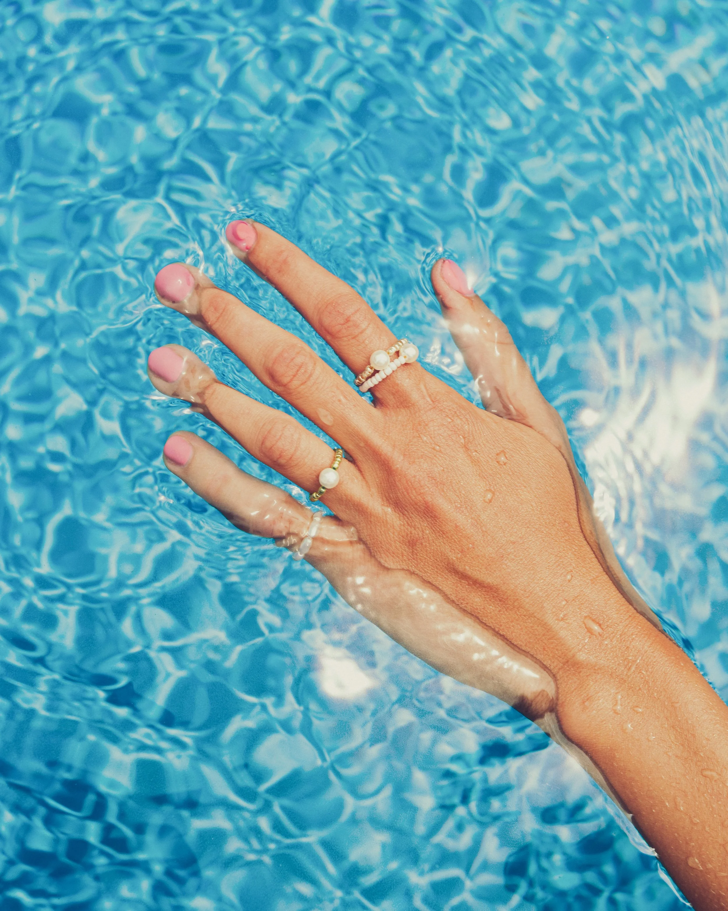 A hand with pink painted nails and multiple rings submerged in clear blue water, creating ripples.
