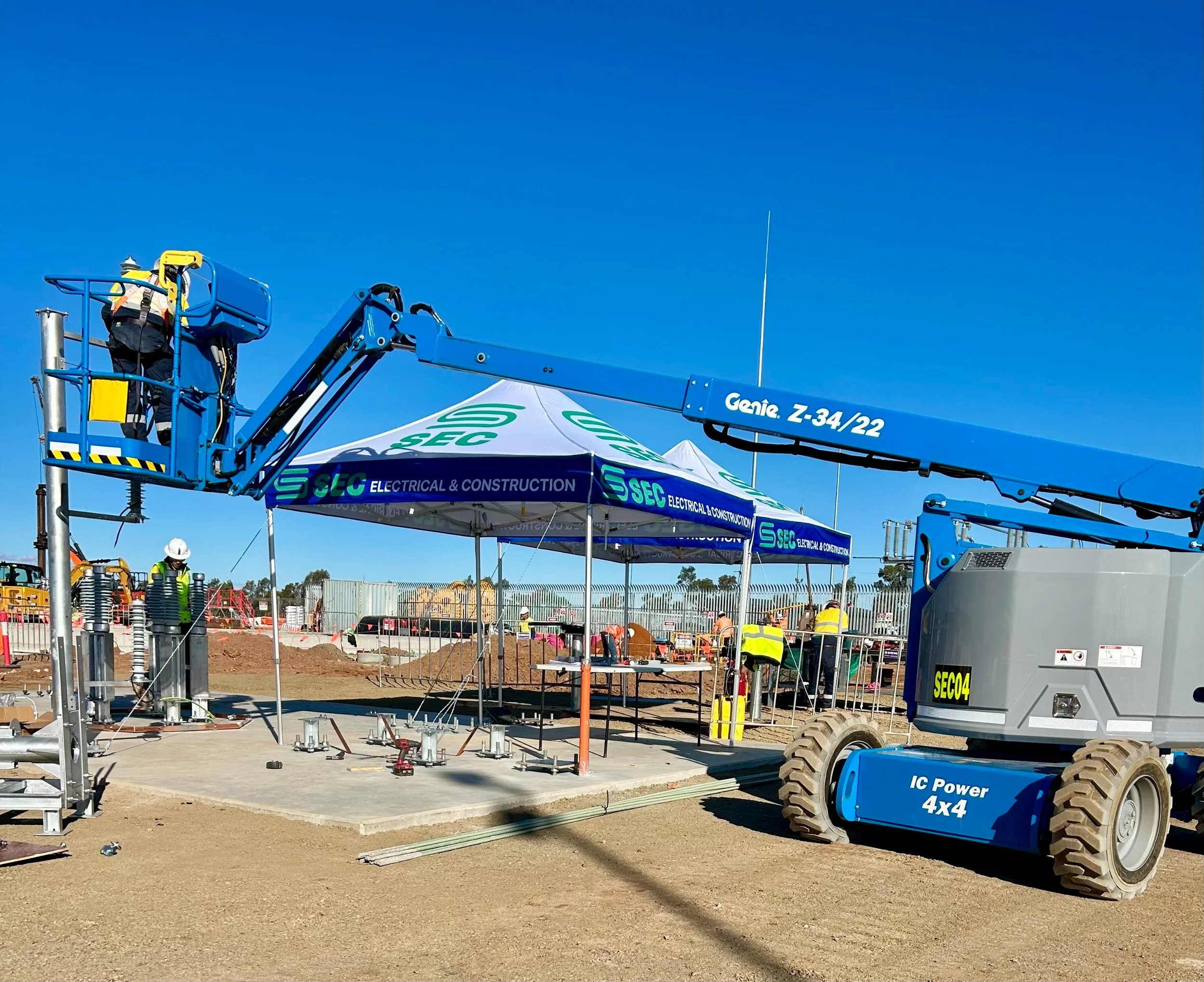 SEC Electrical & Construction team members operating a blue aerial lift at a construction site in Forbes under a clear blue sky, with equipment and materials on the ground.