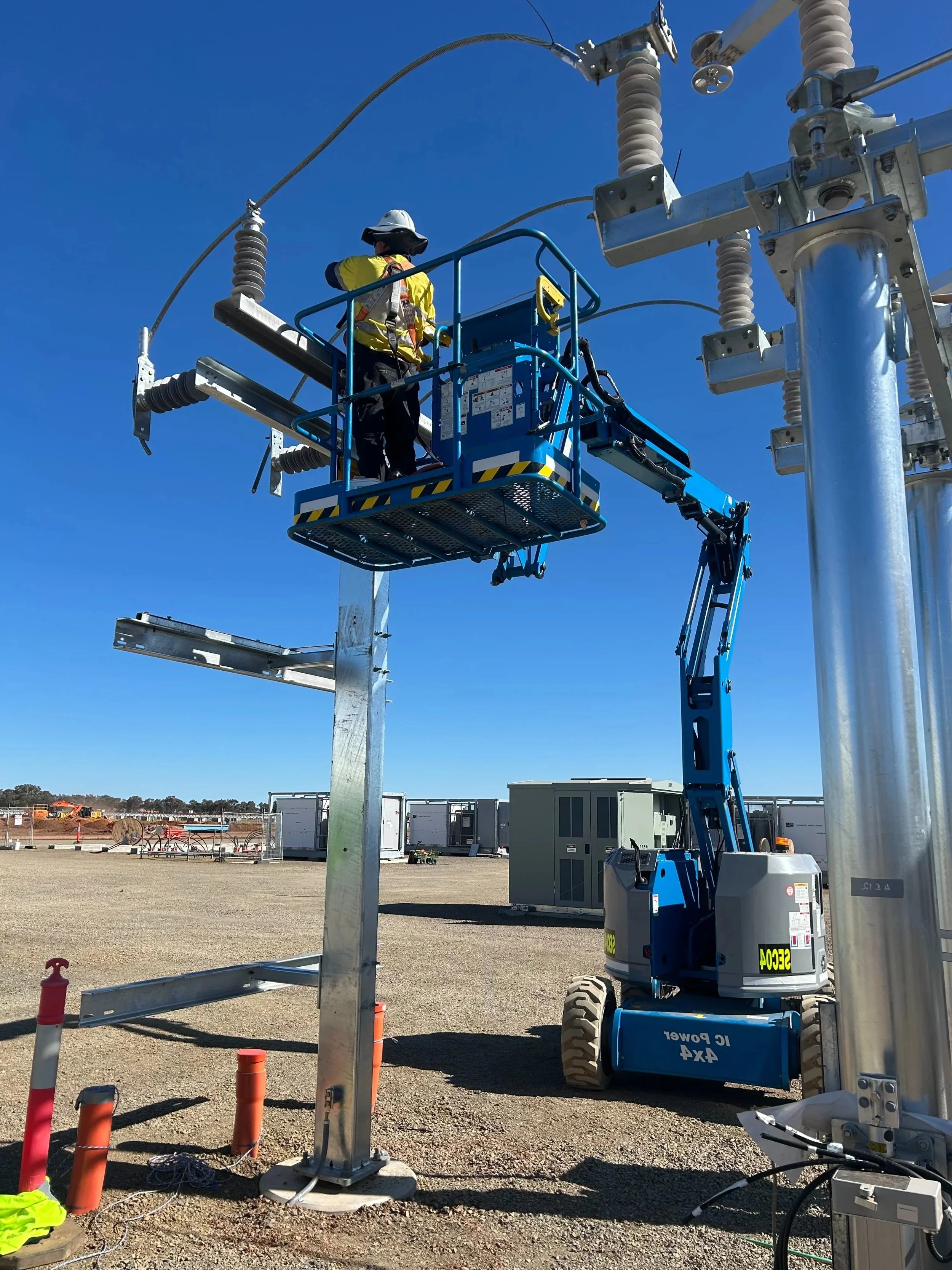 A SEC Electrical & Construction worker wearing safety gear on a blue lift working on an electrical pole in an outdoor construction site with clear blue skies.