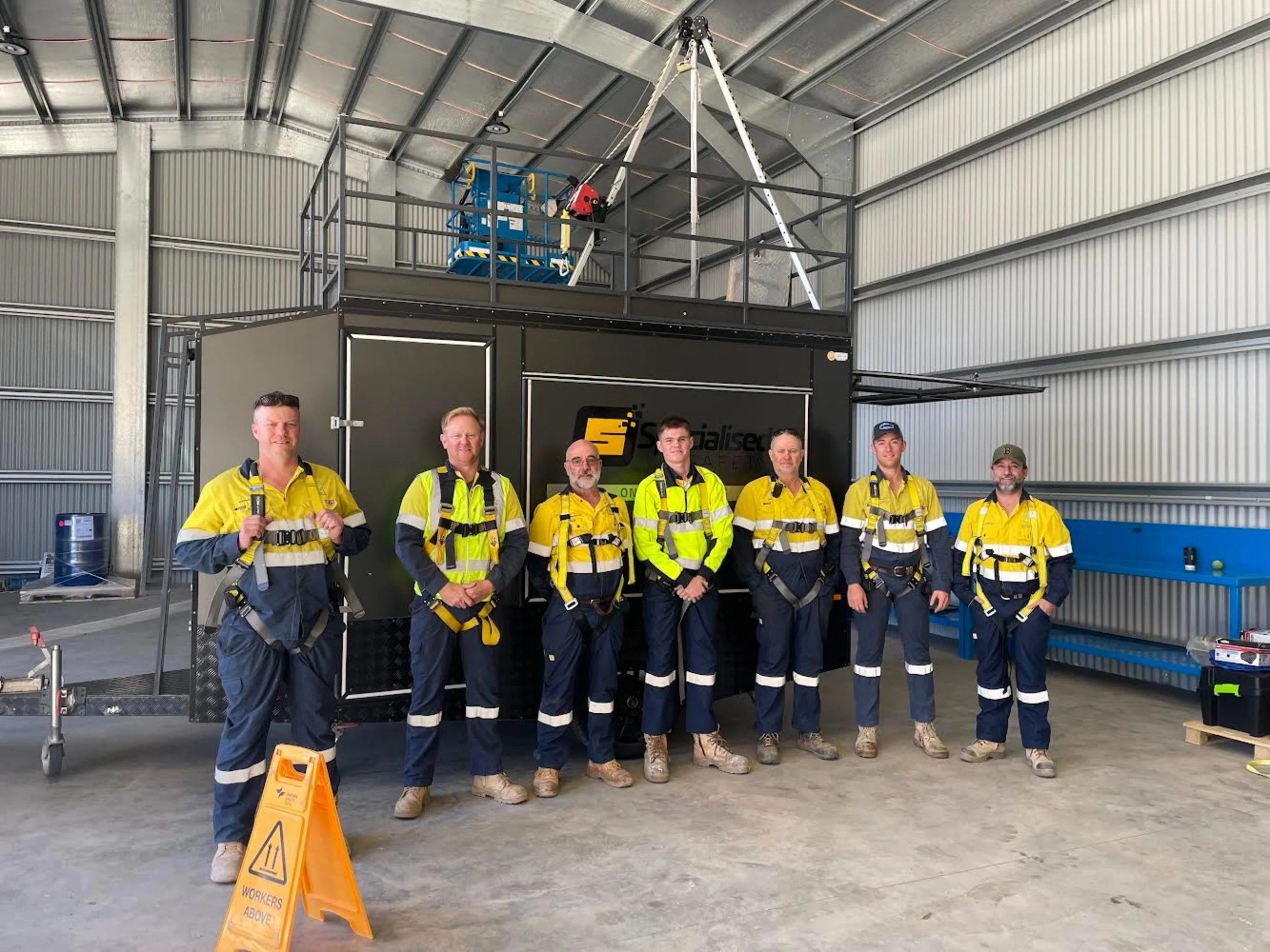 Group of seven SEC Forbes team members in high-vis safety clothing and harnesses standing inside a large warehouse. They are standing in front of a black mobile trailer and behind a yellow caution sign that reads 'WORKERS ABOVE'.