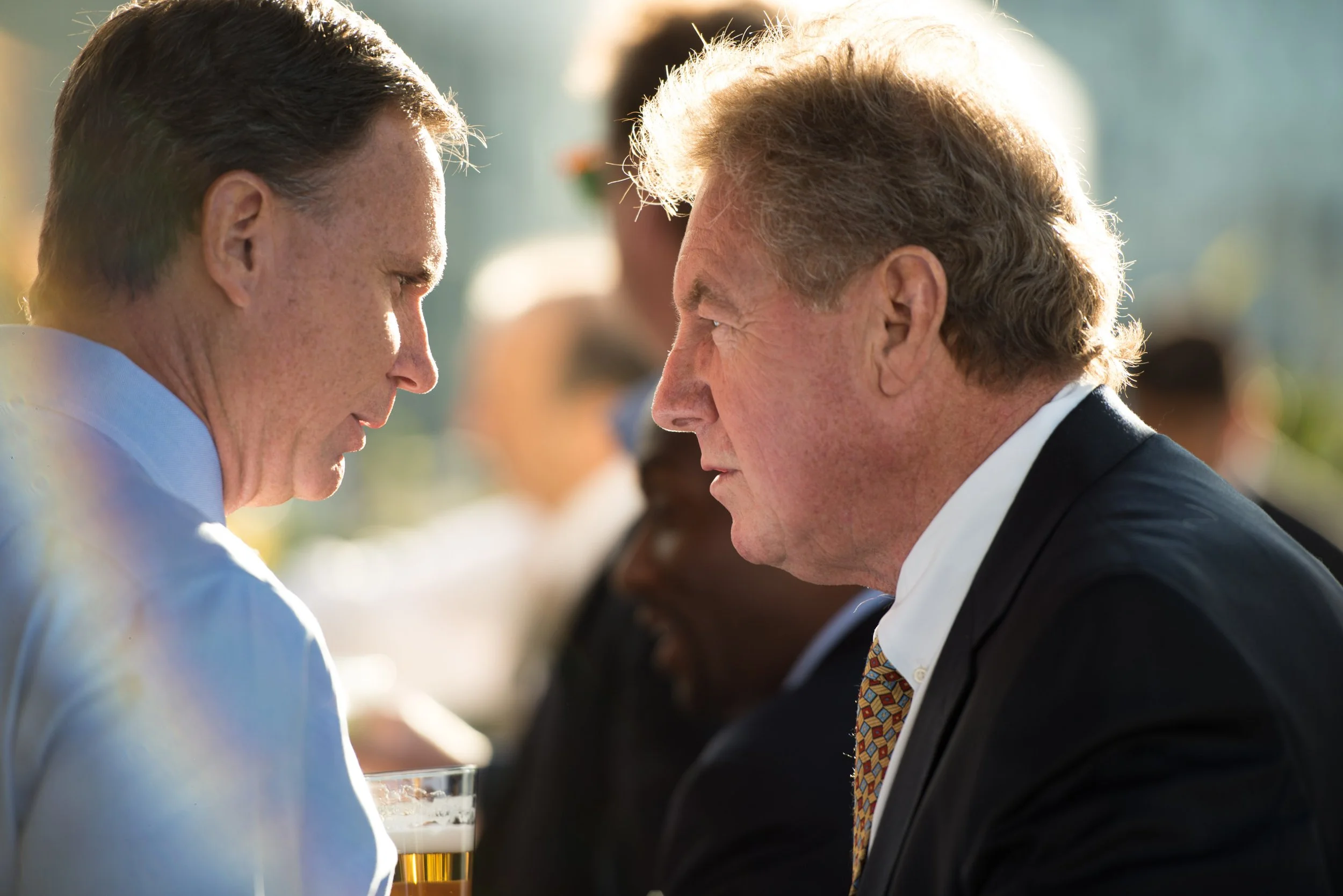 Two men are engaged in a close conversation outdoors, leaning towards each other with serious expressions.