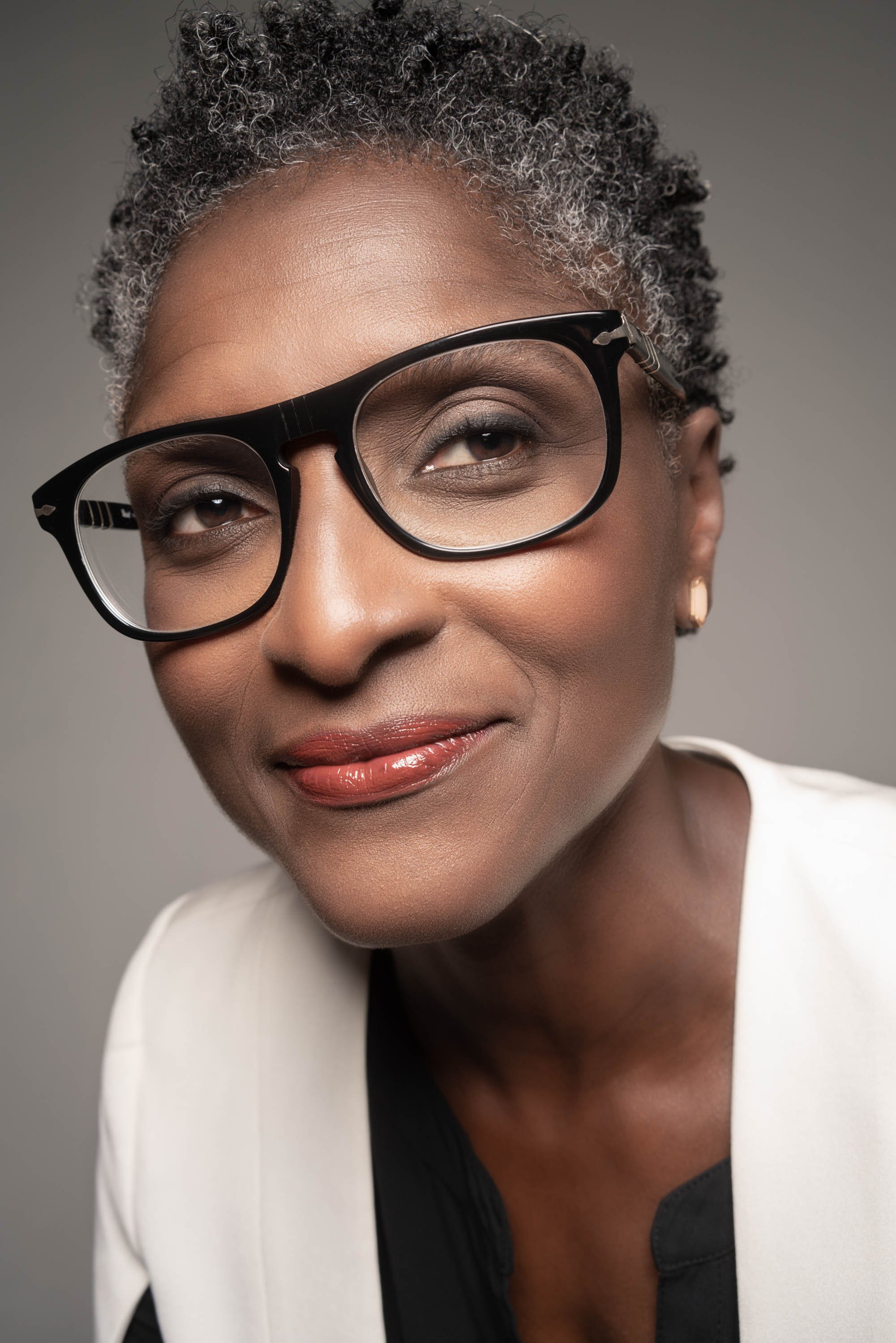 Close-up of an African American woman wearing large black glasses, with short gray curly hair, makeup, earrings, and a white blazer.