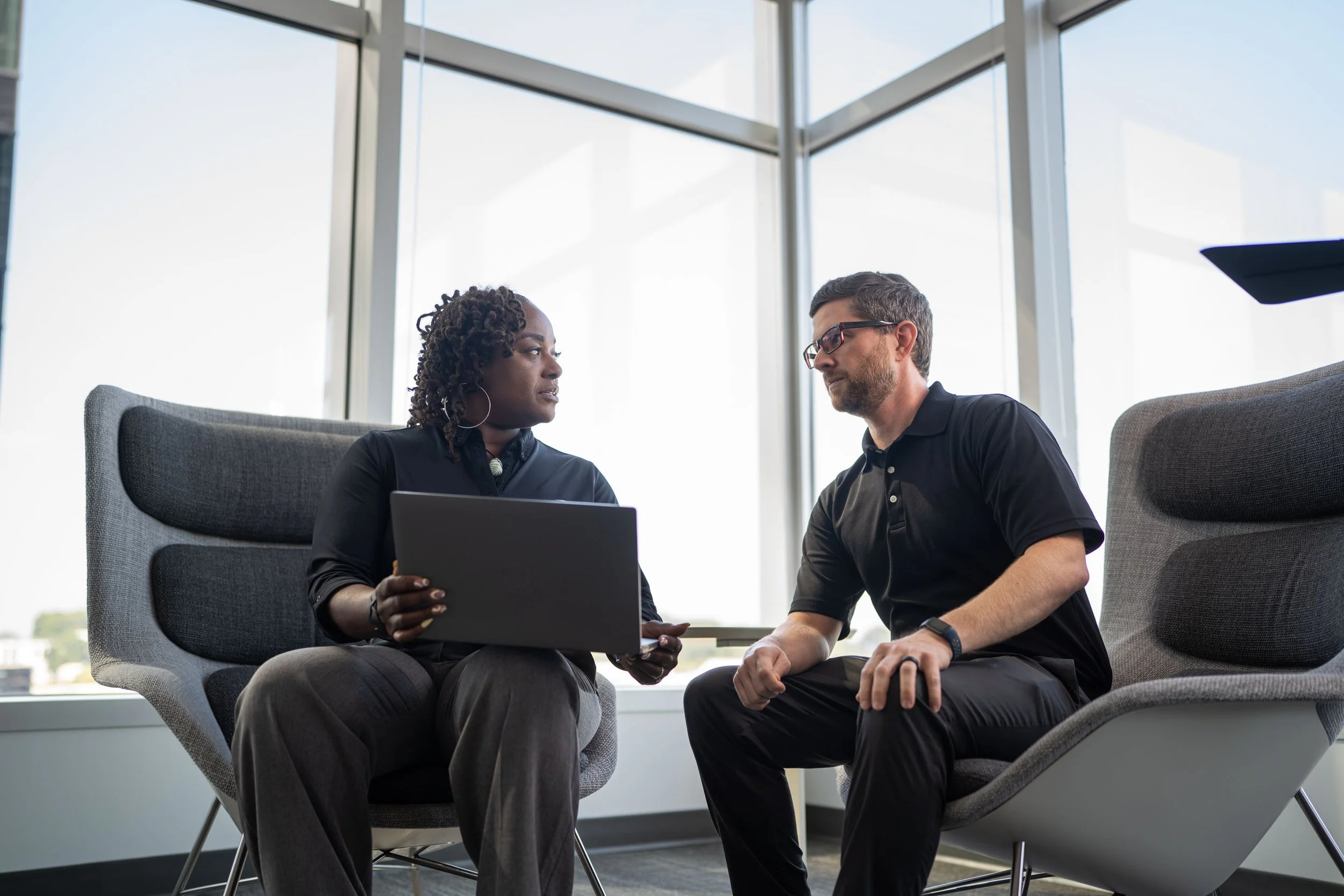 A man and woman are sitting in office chairs facing each other, engaged in conversation, with a laptop in the woman's hands, near large windows in a modern office setting.
