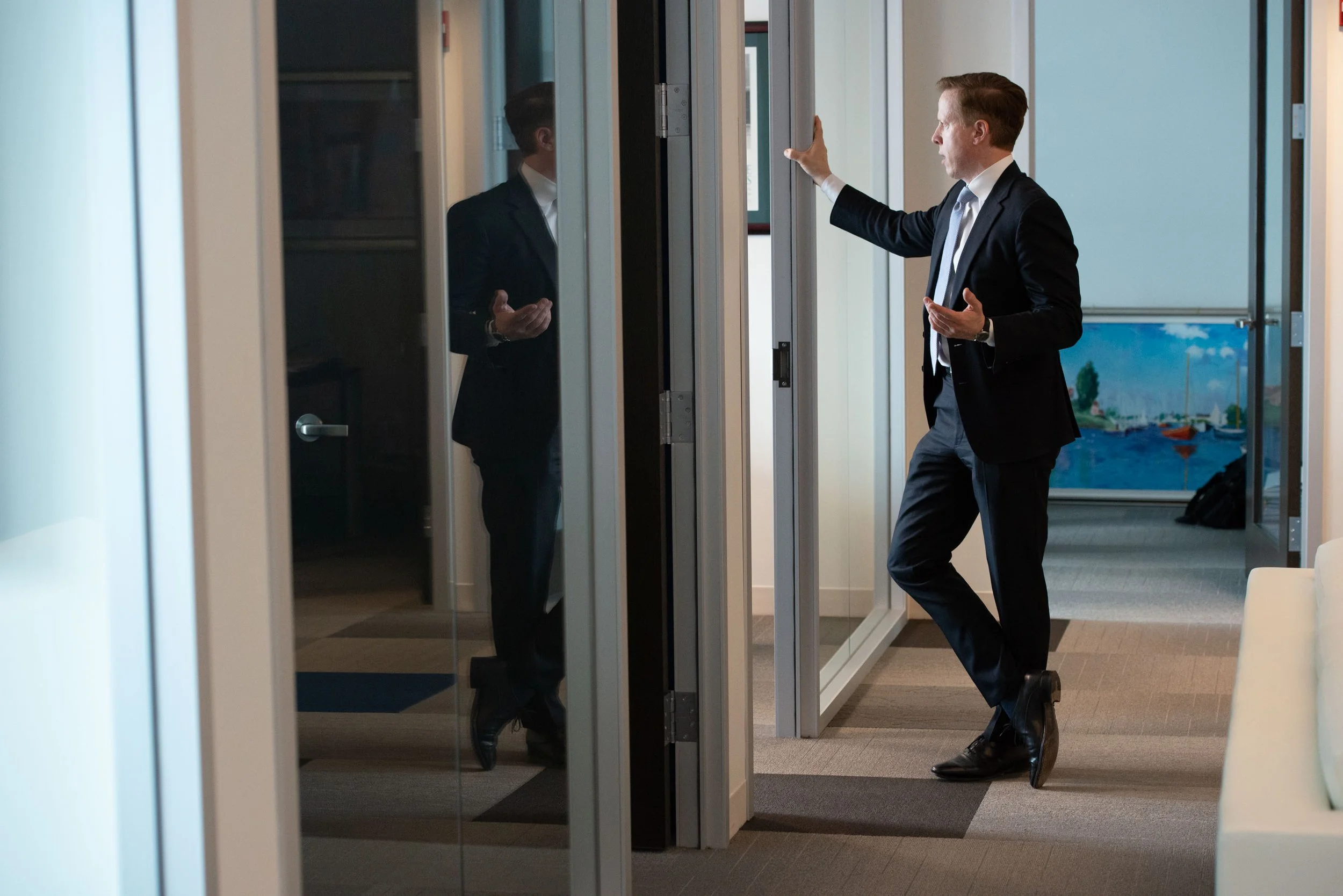 A man dressed in a business suit is talking or presenting, gesturing with his hands, while standing near glass office partitions in a modern office space. His reflection is visible in the glass.