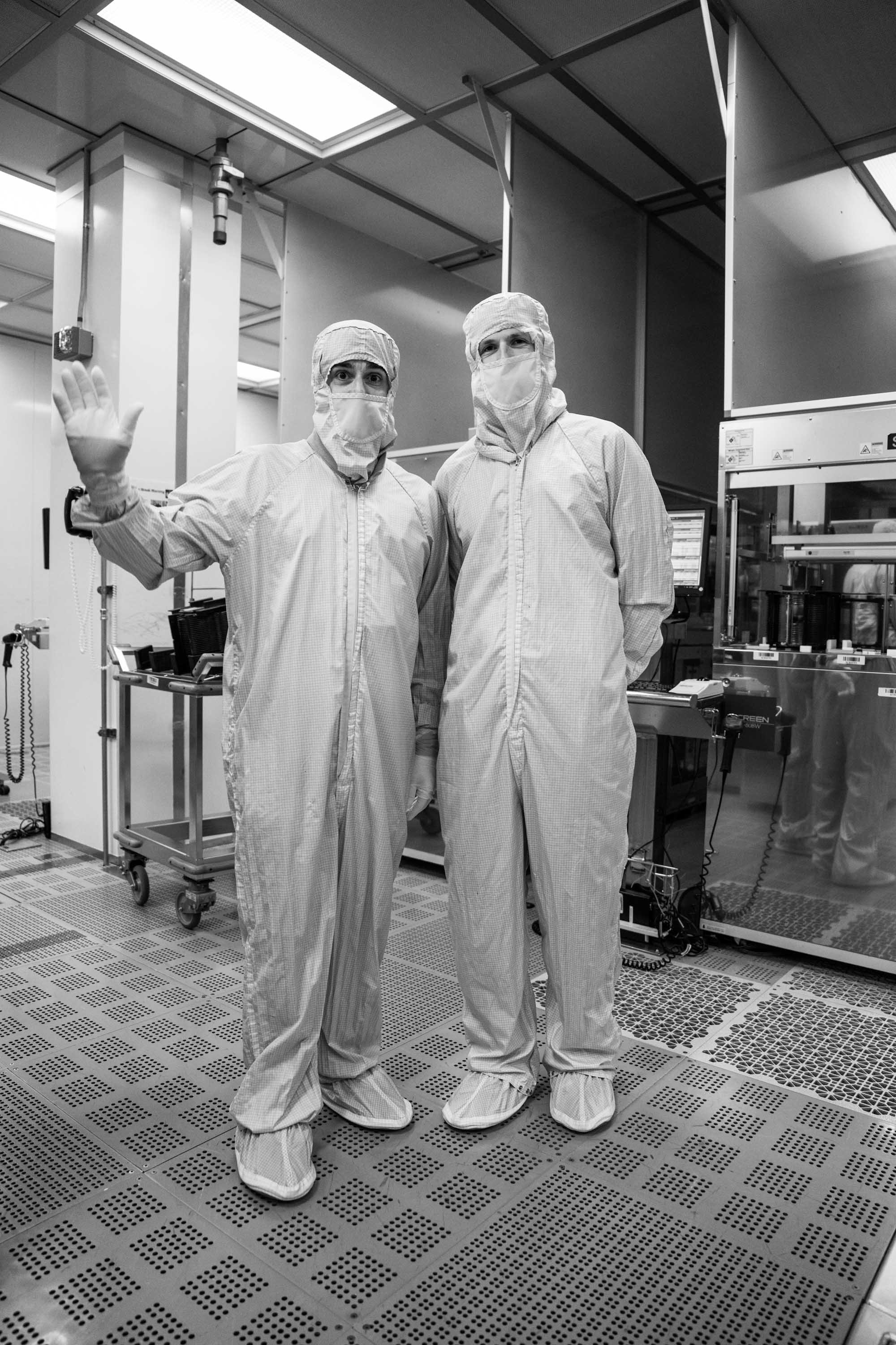Two scientists in full protective suits, masks, and gloves standing in a laboratory. One is waving at the camera. The room contains various scientific equipment and computer monitors.