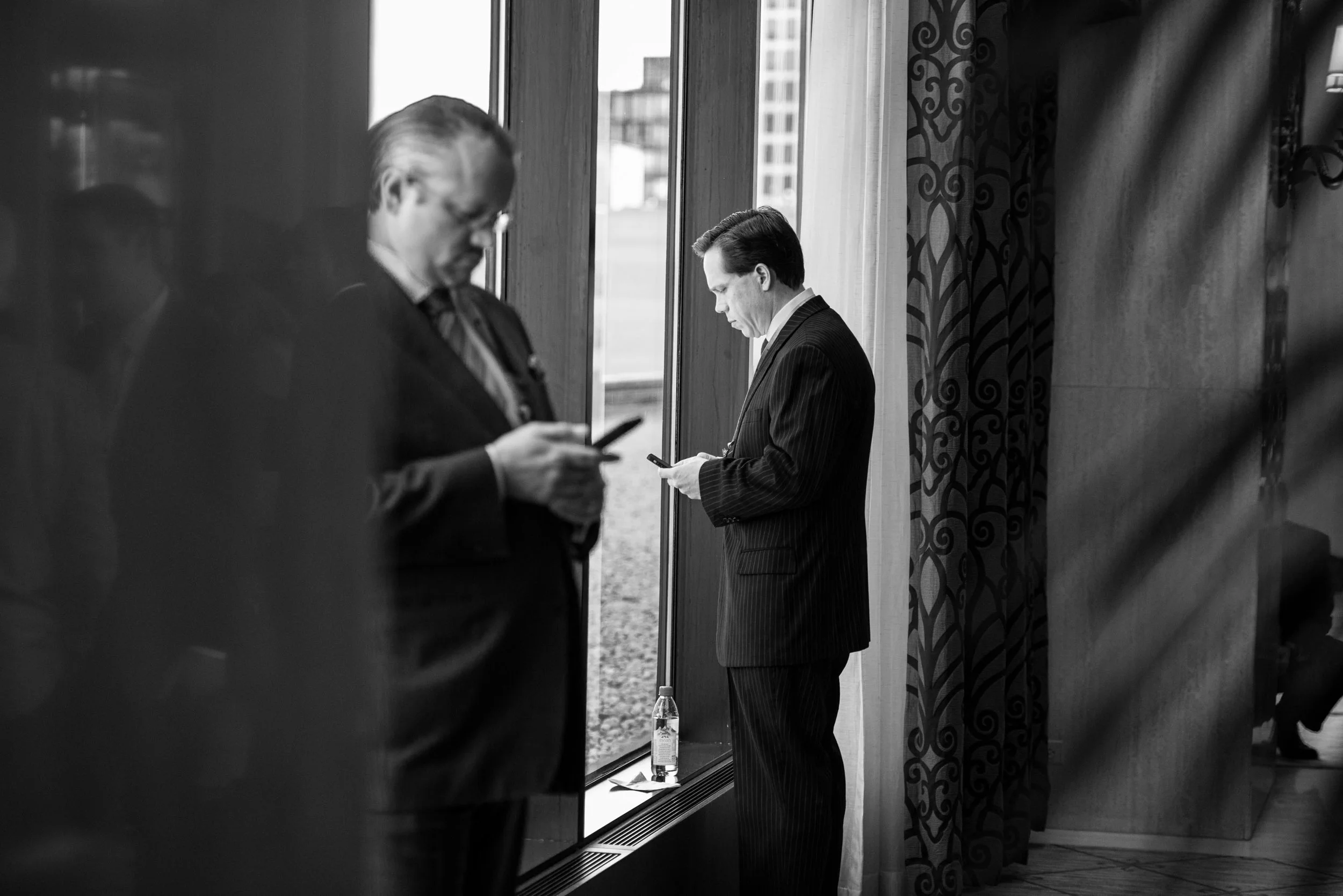 Two suited male executives checking smartphones by a large window during a corporate conference break, city skyline visible, black and white — Los Angeles corporate event photographer — Sightline Visuals